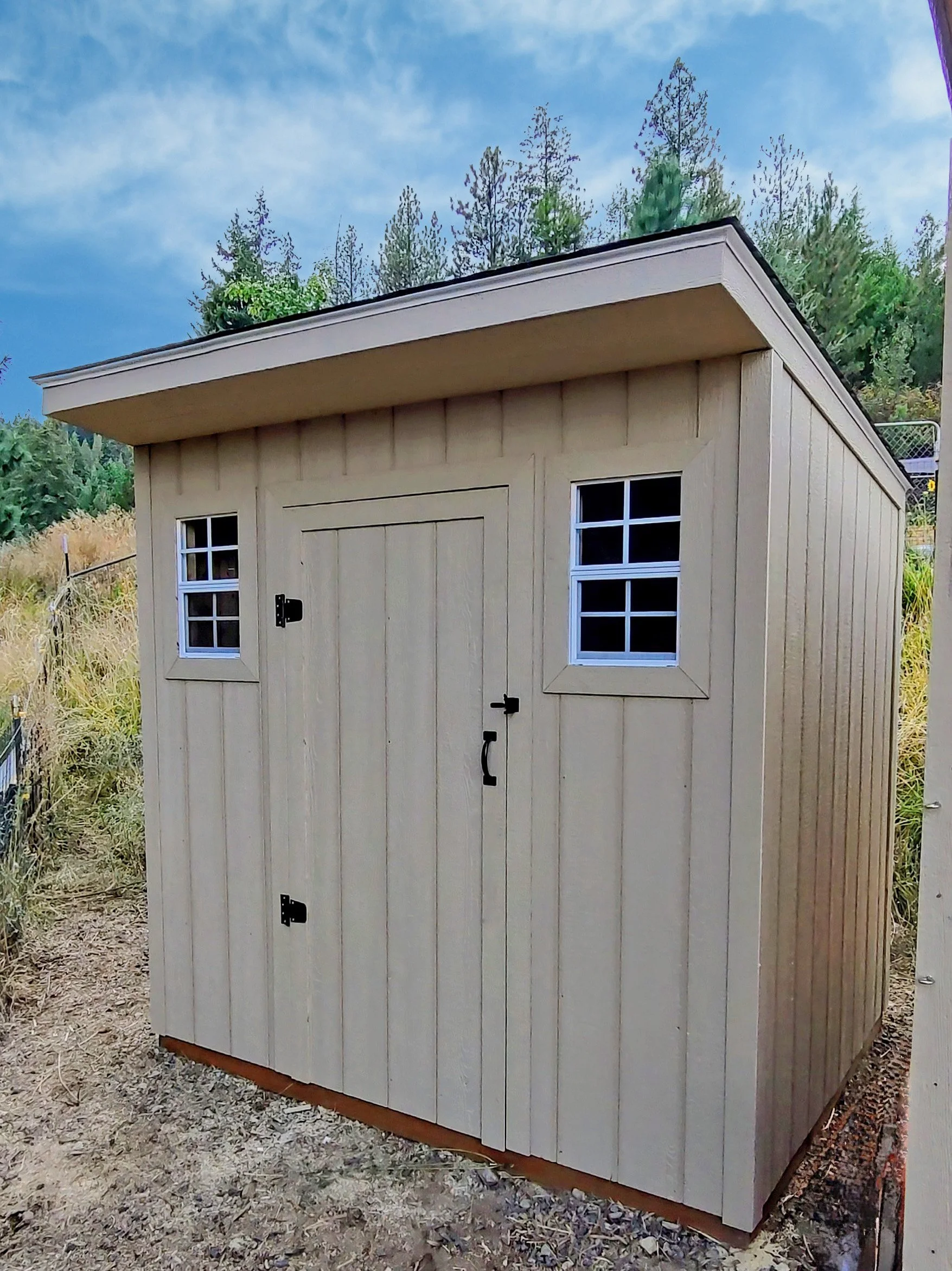A small beige wooden shed with two small windows and a door, located outdoors with trees and a partly cloudy sky in the background.