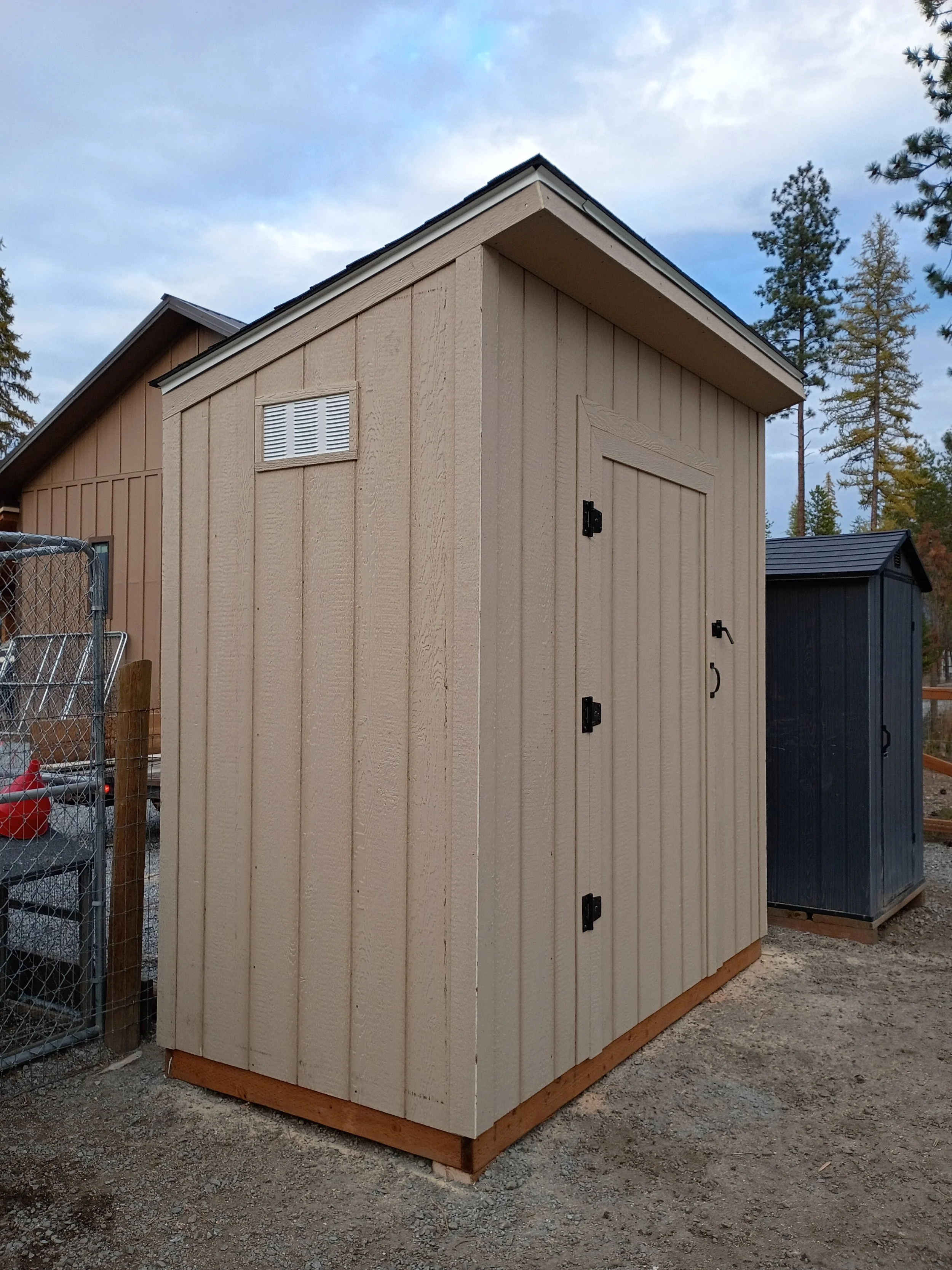 A beige wooden outdoor shed with black hinges and a door, situated on a gravel surface in a backyard with trees in the background.