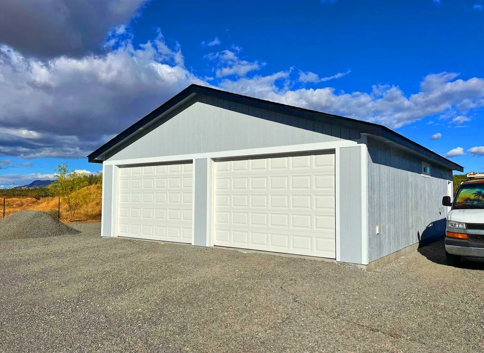 A white garage with two closed garage doors situated on a gravel lot under a partly cloudy sky.
