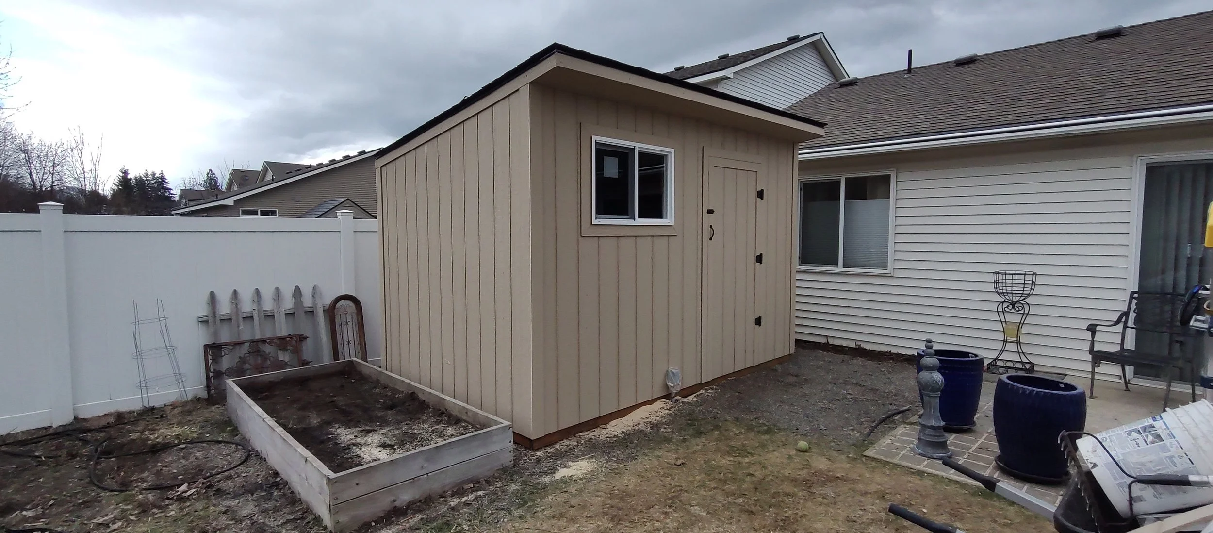 Backyard with beige shed, window, white vinyl fence, garden bed, outdoor chair, potted plants, and garden decorations under cloudy sky.