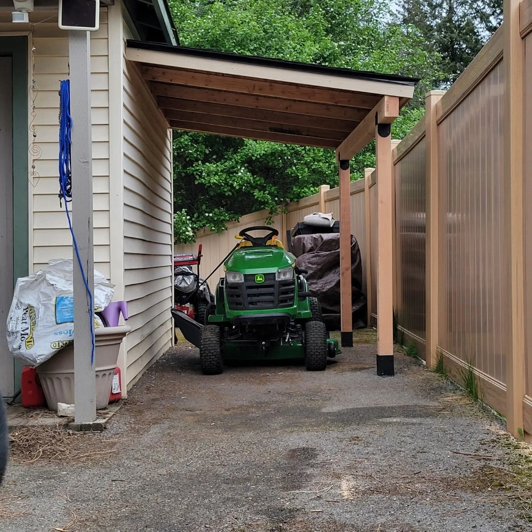A small backyard storage area with a green John Deere riding lawn mower, covered outdoor furniture, and a shed with a wooden roof cover. There is a beige vinyl fence on the right side and a beige house with white siding on the left. Garden tools and supplies are partially visible.