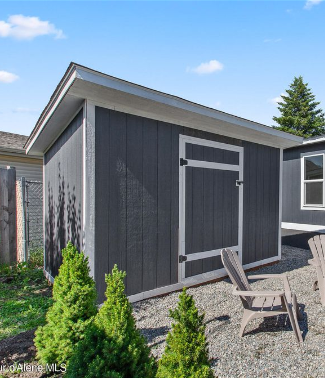 A dark gray wooden outdoor shed with white trim, set on gravel, with two Adirondack chairs and small green bushes in the foreground.