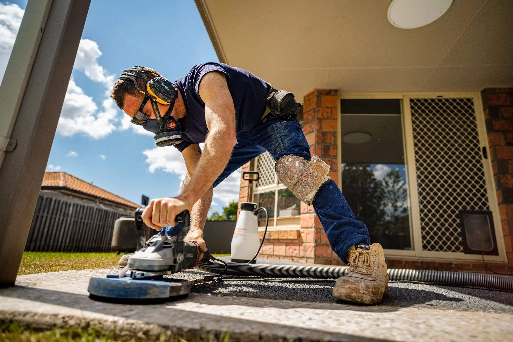 A man wearing safety gear, including ear protection, a face mask, and knee pads, is using a grinder on a concrete surface outside a house with brick walls and a screened door.