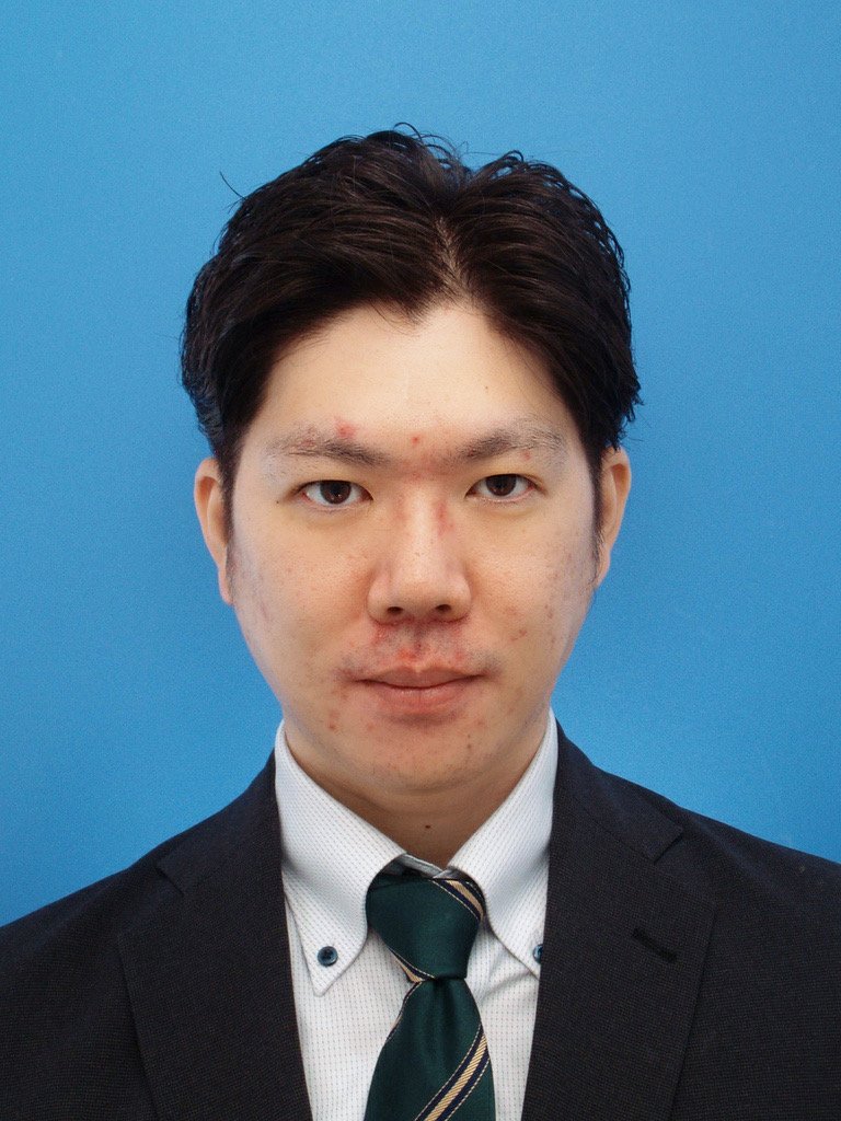 青い背景の前に立つ若い男性の肖像写真。白いシャツにネクタイ、黒いスーツを着ている。