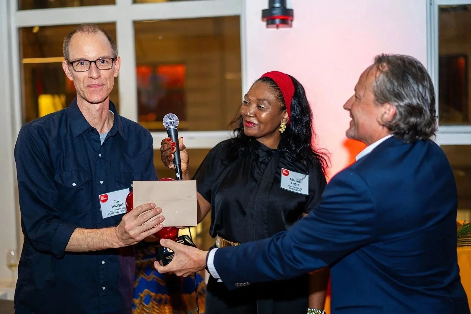 Three people at an awards event: a man with glasses holds a plaque, a woman with a red headband and earrings looks on, and another man in a blue suit hands over the plaque, smiling.