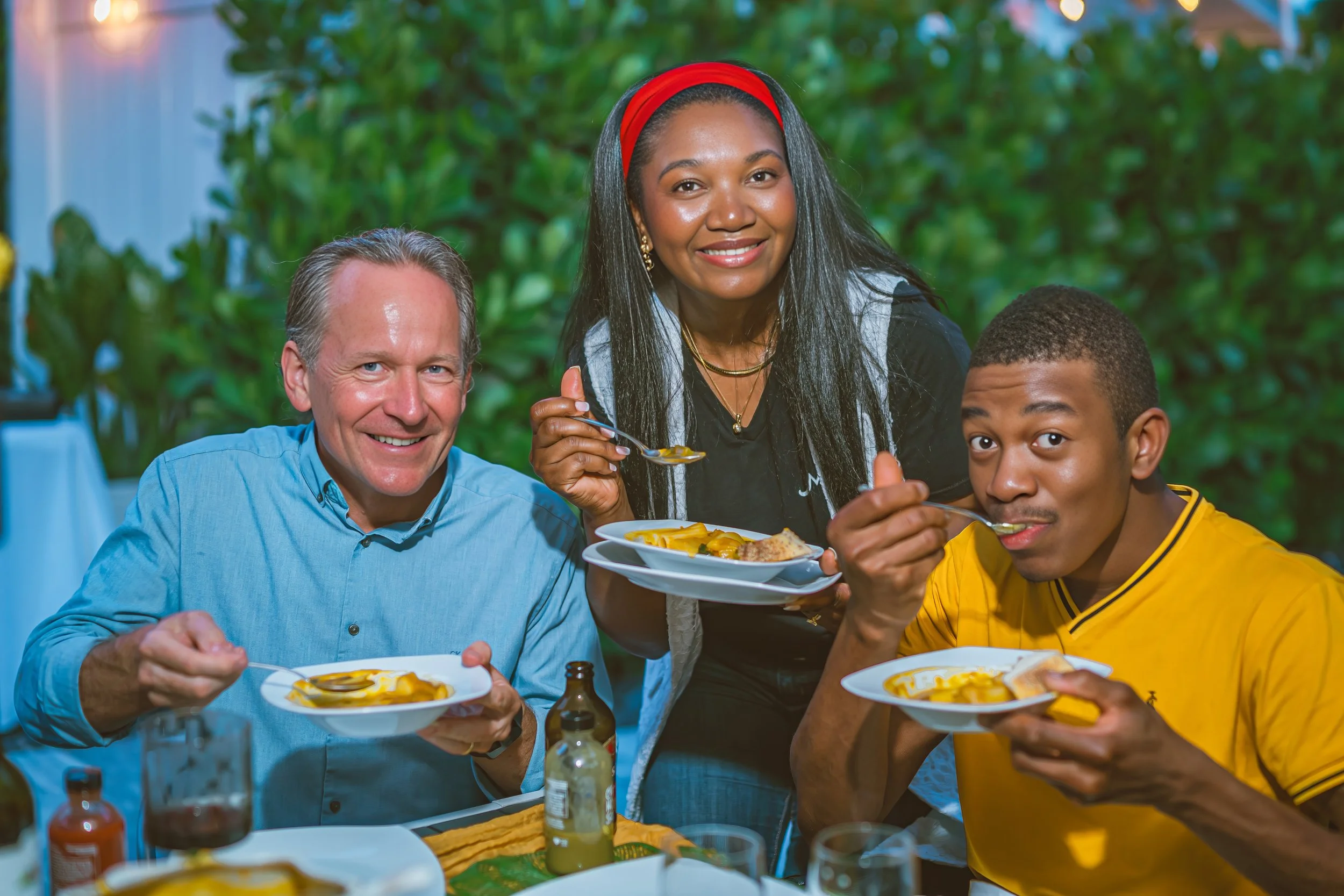 Three people enjoying a meal at a table outdoors, with two men and a woman smiling while eating yellow pasta and meat