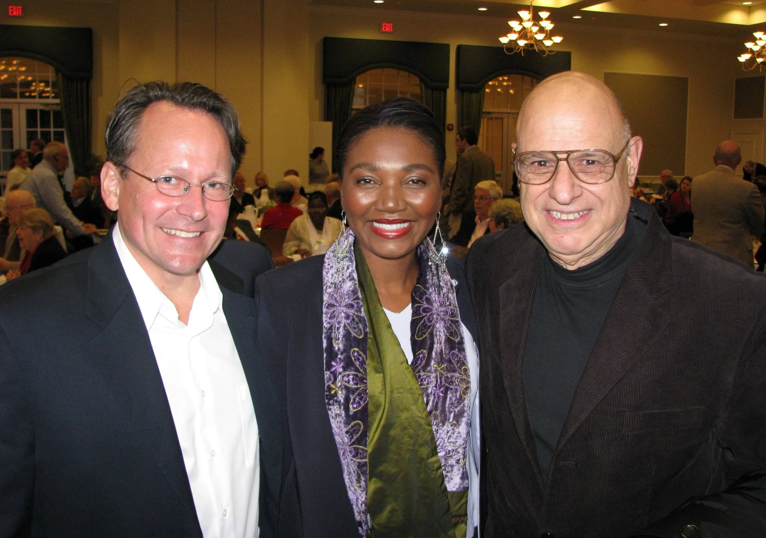 Three people, two men and one woman, smiling and posing for a photo at an indoor event with many attendees seated at tables in the background.