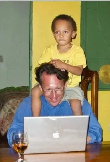 A young girl sitting on an adult man's shoulders, using a laptop at a table with a glass of drink nearby.