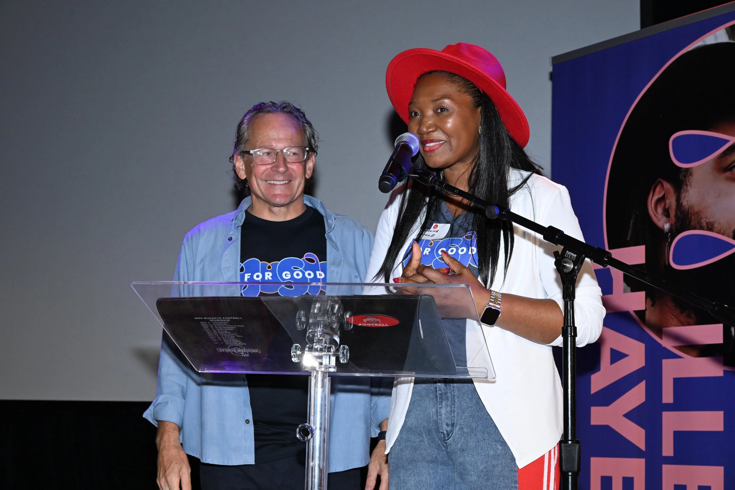 Two women standing at a podium on stage, speaking into a microphone. One woman has long black hair, wearing a red hat, white blazer, jeans, and earrings. The other woman has short gray hair, glasses, and is wearing a black T-shirt and light blue shirt. Both women are smiling, and the background features a large banner with part of a face and the words "FOR GOOD."