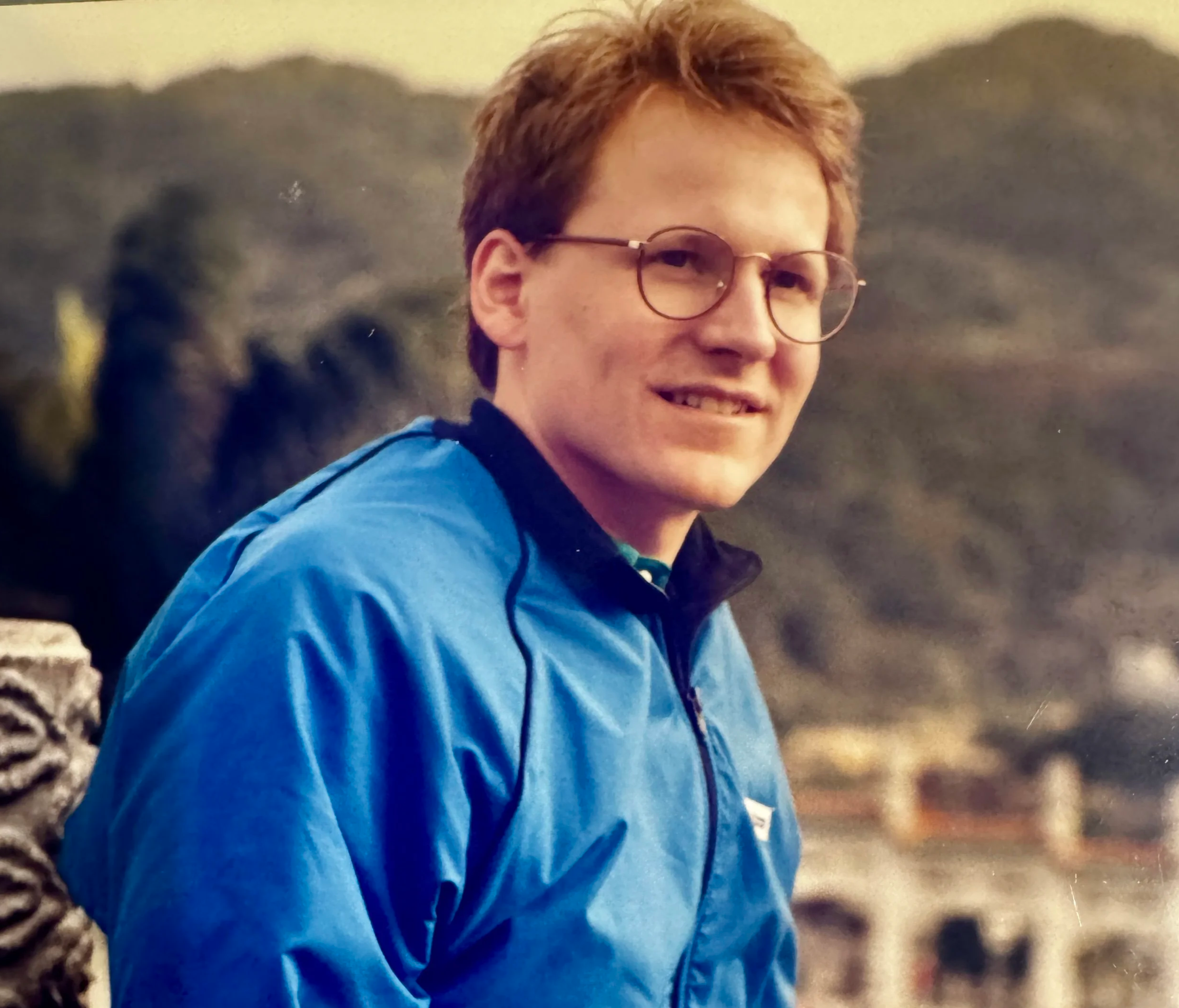 A person with short reddish-brown hair and glasses, wearing a bright blue jacket, smiling at the camera with a blurred European cityscape in the background.