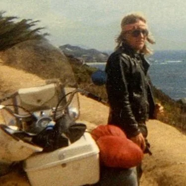A person with blonde hair wearing sunglasses and a black leather jacket standing beside a motorcycle on a coastal trail.