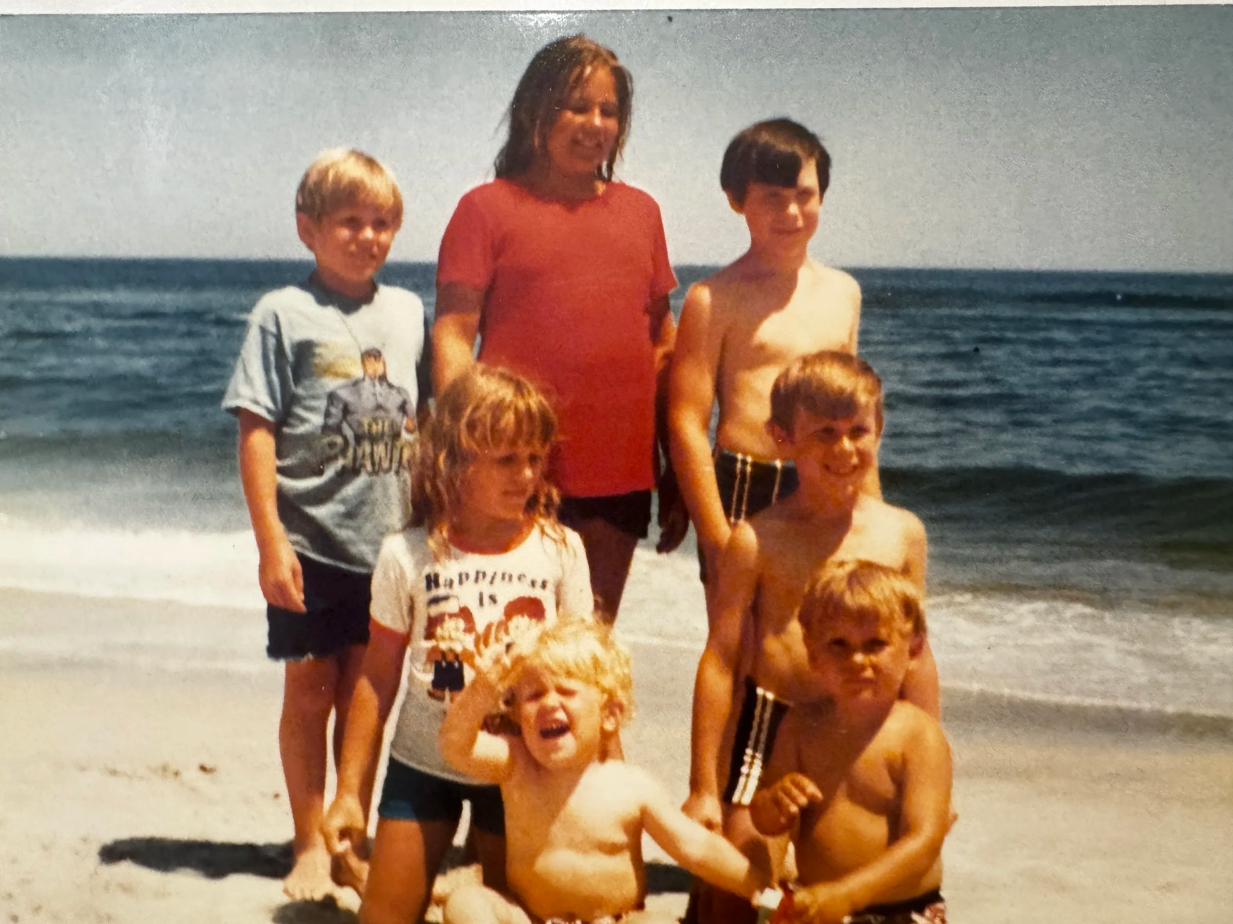 A group of seven children and one adult at the beach, posing for a photo with ocean waves in the background. The children are wearing various casual clothes and swimwear, some smiling and some making funny faces.