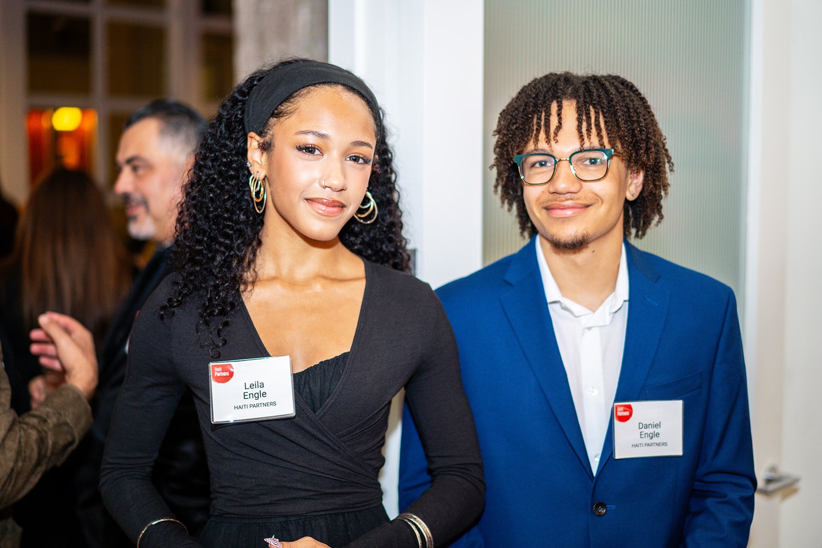 Two young adults, a woman and a man, wearing name tags at a professional event. The woman has curly dark hair, earrings, and is dressed in black. The man has curly hair, glasses, and is wearing a blue suit. Both are smiling.