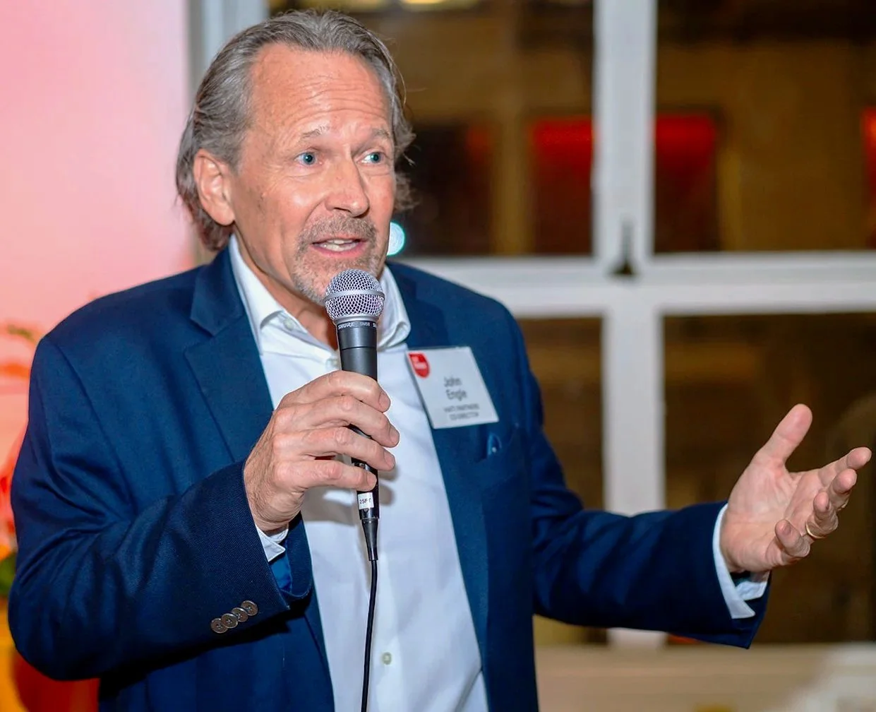 A man in a blue suit and white shirt is speaking into a microphone, gesturing with his left hand, during an indoor event.