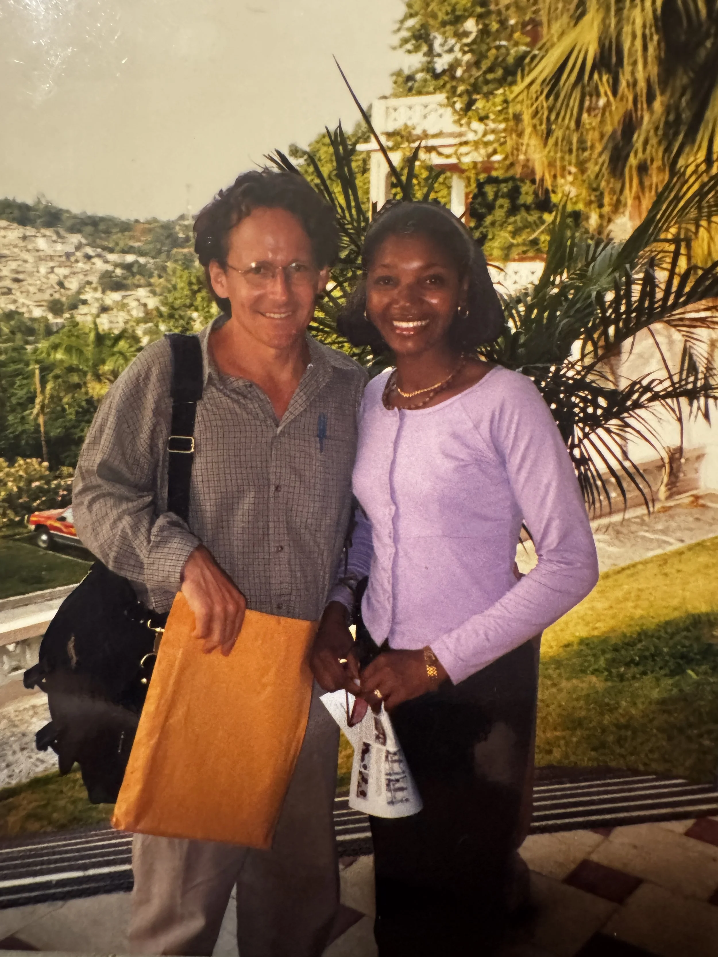 A man and a woman standing together outdoors with greenery and trees behind them, smiling at the camera.
