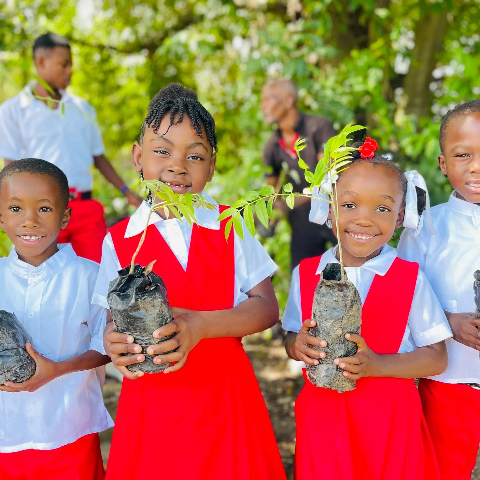 Group of children in school uniforms holding small potted trees outdoors, smiling, with greenery in background.