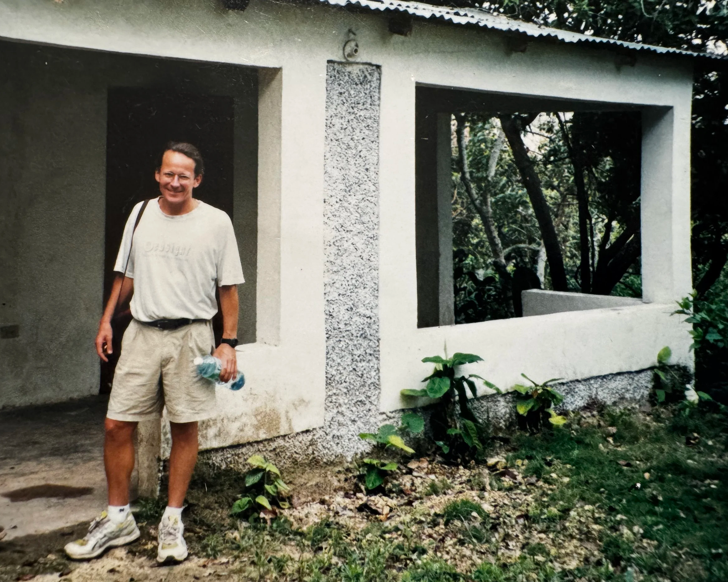 A man smiling, standing outside a partially constructed or renovated building, holding a water bottle, with surrounding greenery.