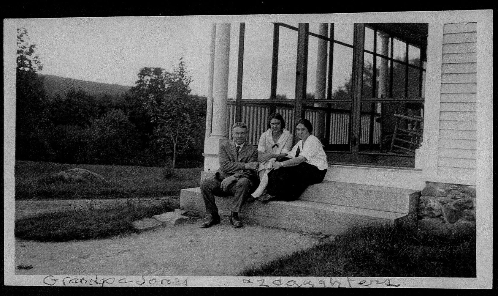 Black and white photo of three people sitting on steps of Wolf & Harp, with a scenic outdoor background of trees and hills of Norway Maine
