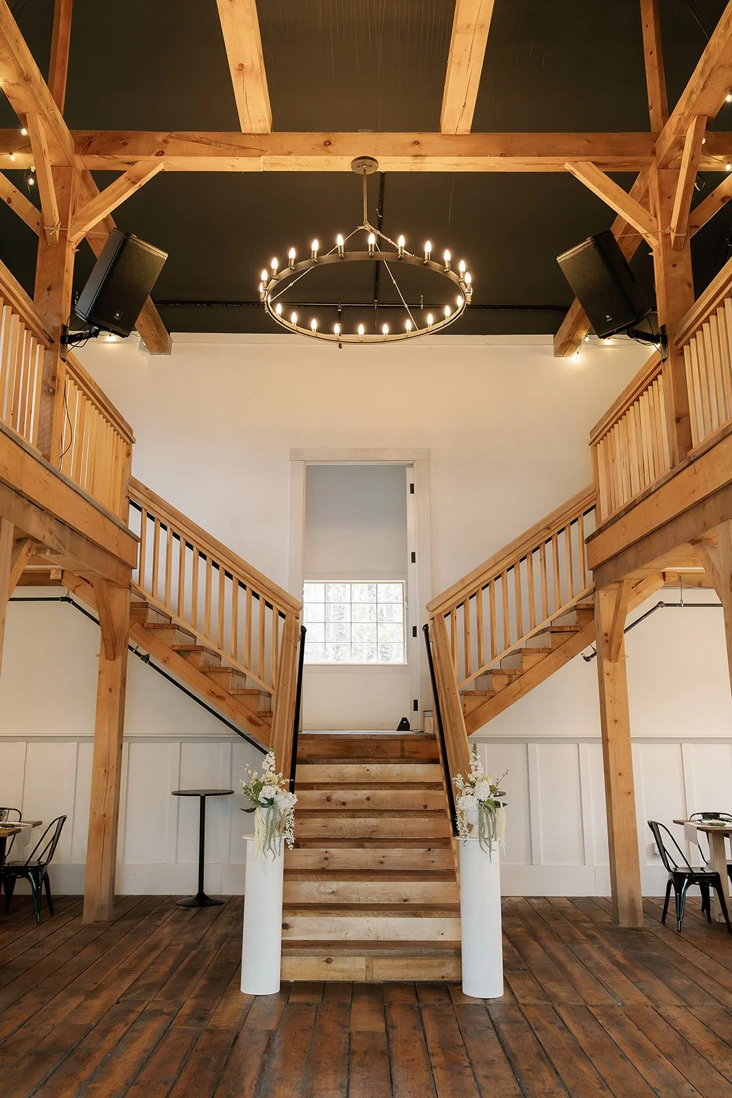 Interior of a rustic venue with wooden staircase leading to an upper balcony, chandelier hanging from a black ceiling, and speakers mounted on the ceiling, with tables and chairs visible on sides.