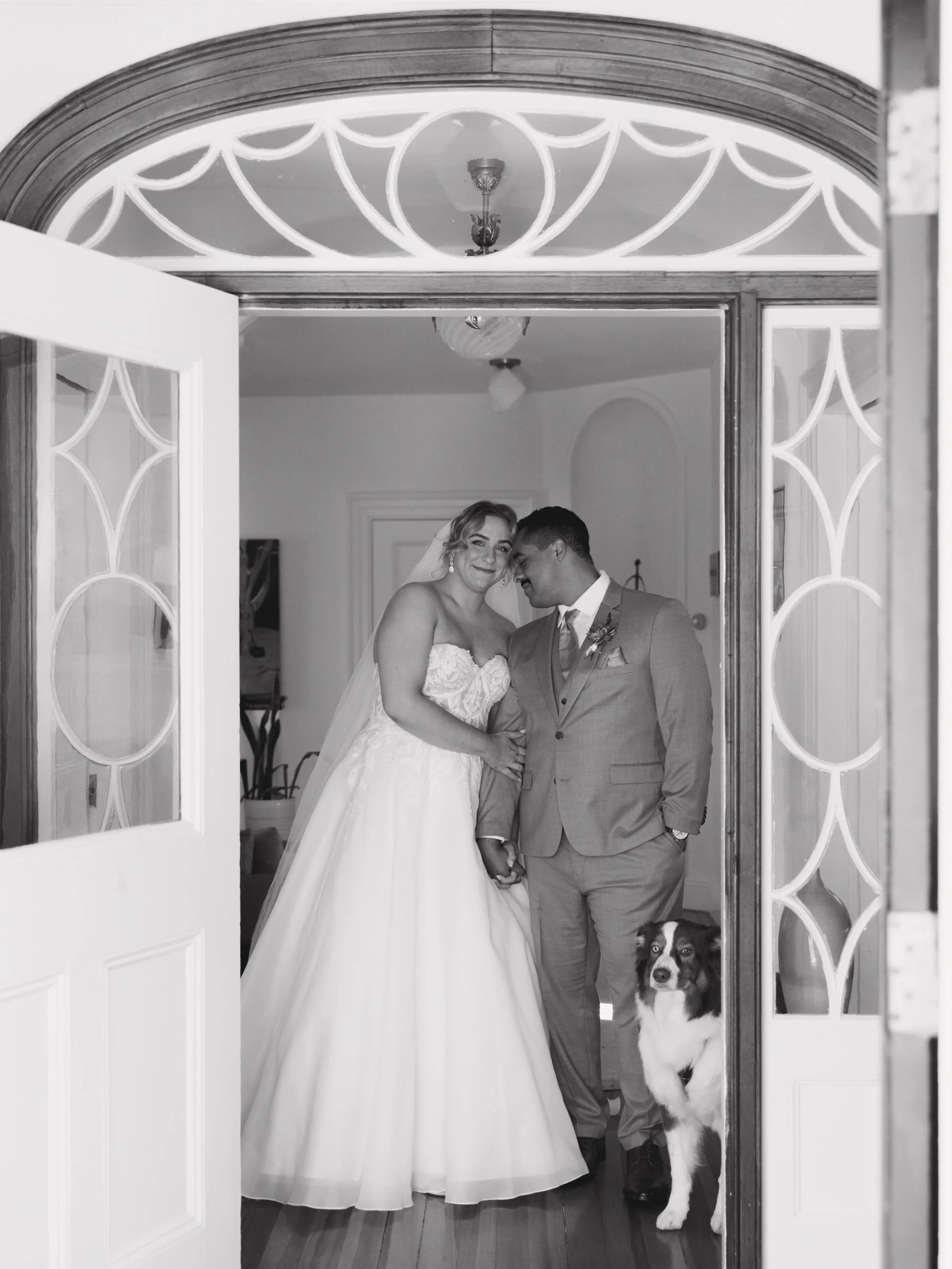 Black and white photo of a bride and groom smiling and holding hands inside a doorway, with a dog sitting on the floor next to them.