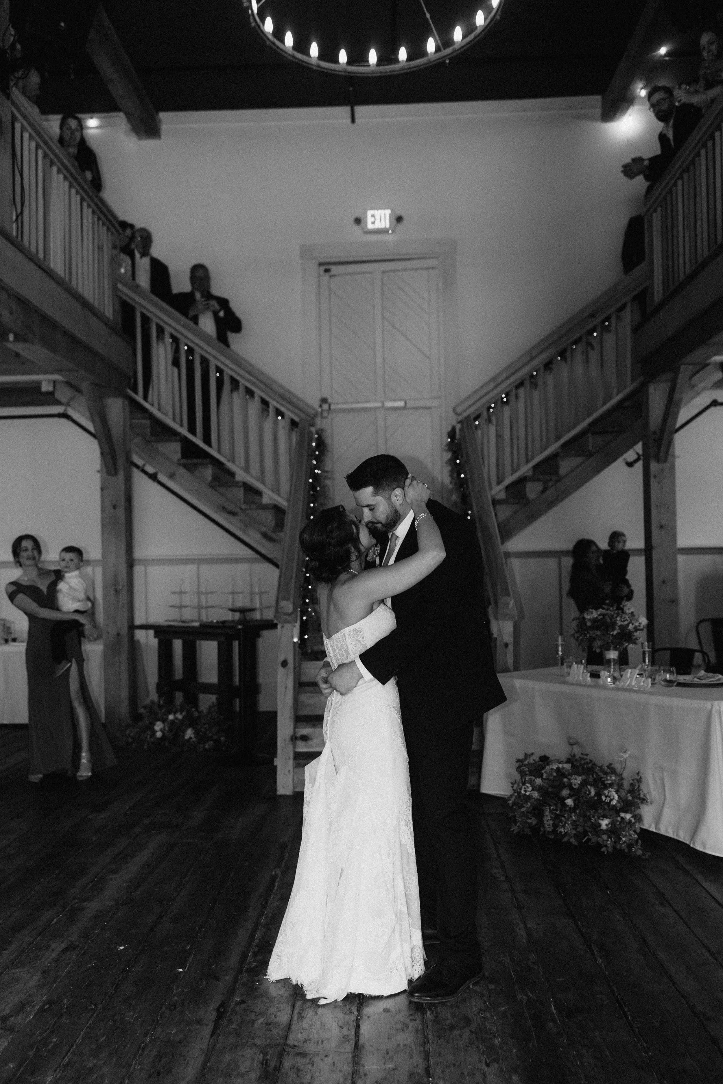 A black-and-white photo of a bride and groom dancing at their wedding reception in a rustic venue with wooden floors and staircases. Guests are watching from the upper level and in the background.