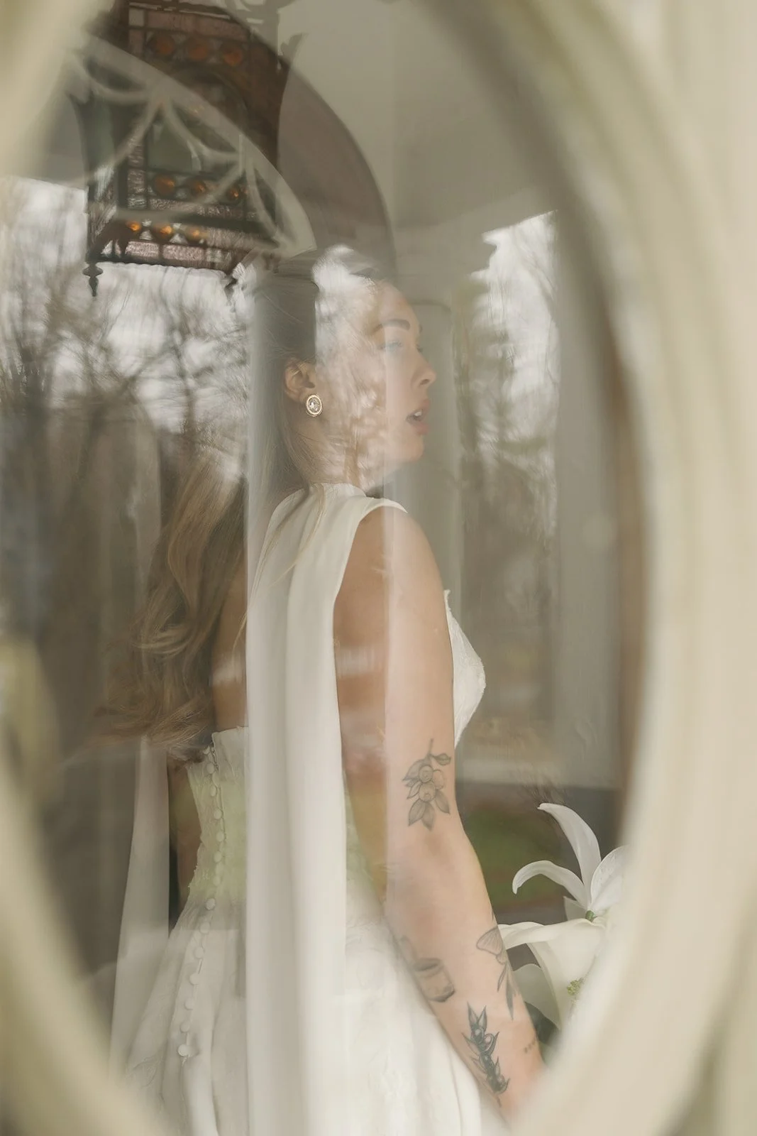 A woman in a white dress with tattoos on her arm, looking to the side through a window with reflections of trees and a hanging lantern.