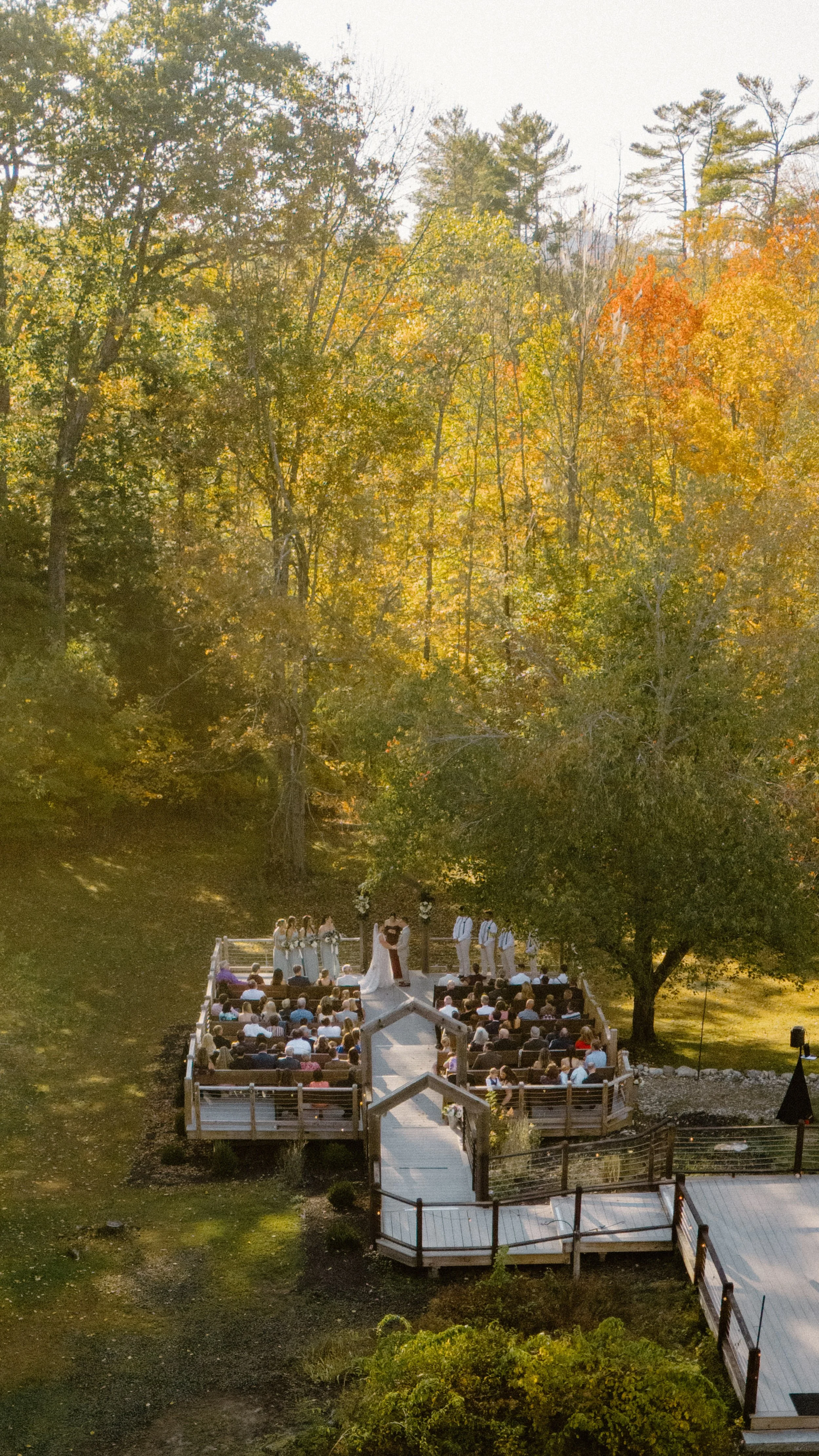 Outdoor wedding ceremony taking place on a wooden platform surrounded by trees with autumn leaves, with guests seated attending the service.