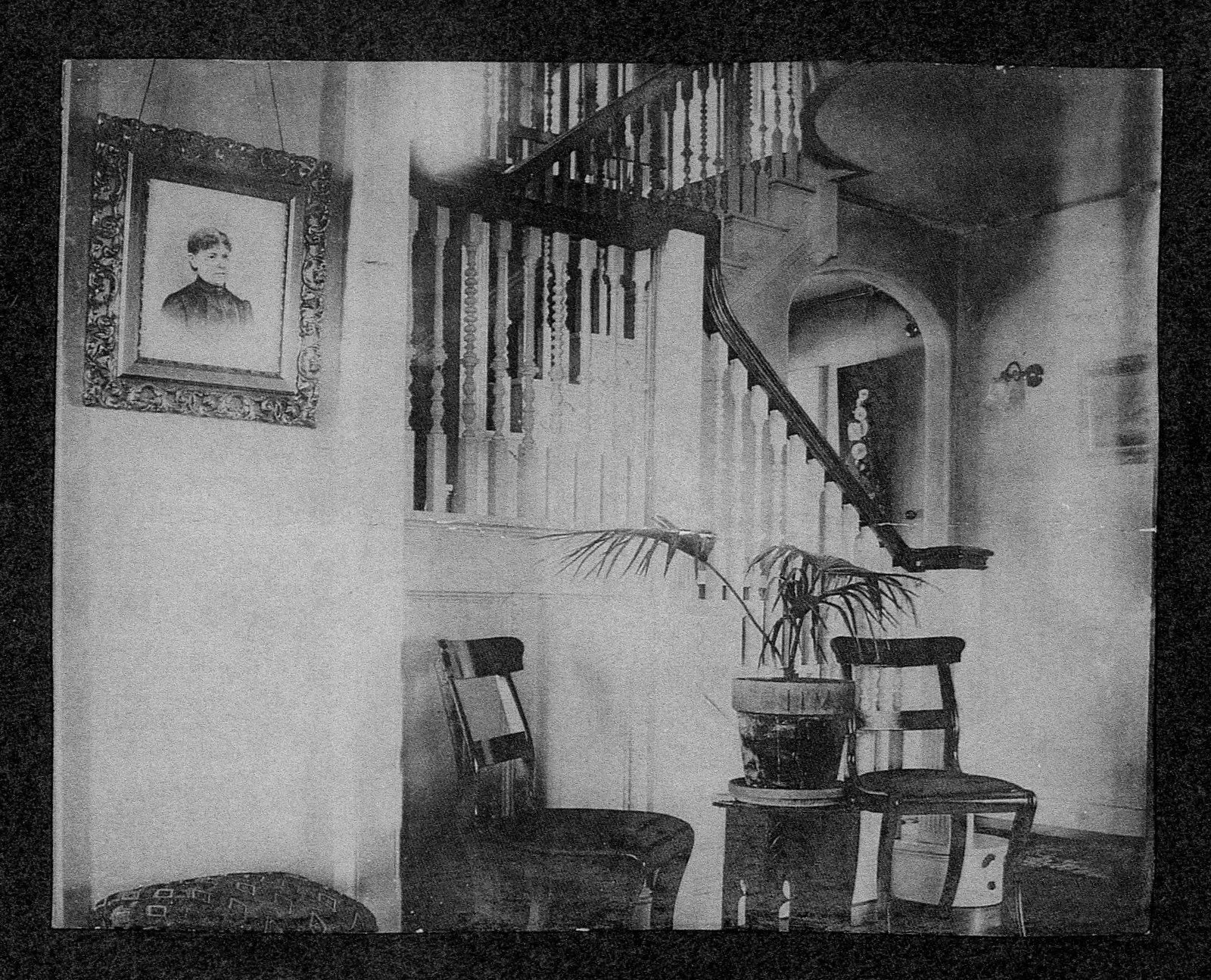 Interior of a house with two wooden chairs, a potted plant, a staircase, and a portrait portrait on the wall.