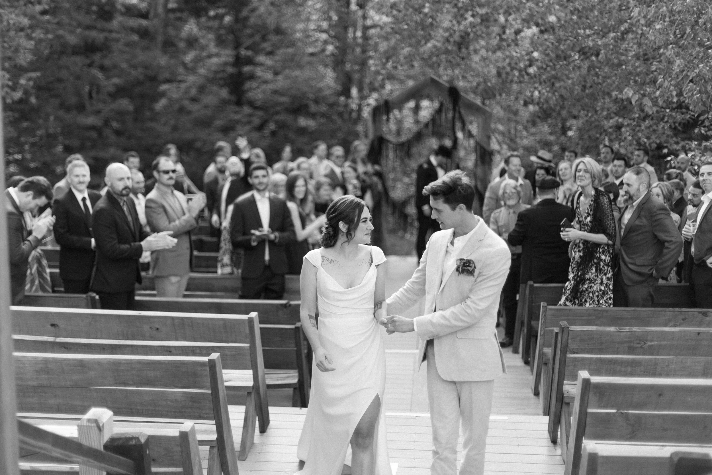 A black-and-white photo of awedding ceremony outdoors with the bride and groom standing close, surrounded by guests in formal attire, seated on wooden benches. The couple appears to be exchanging vows or rings, with trees and a decorated arch in the background.