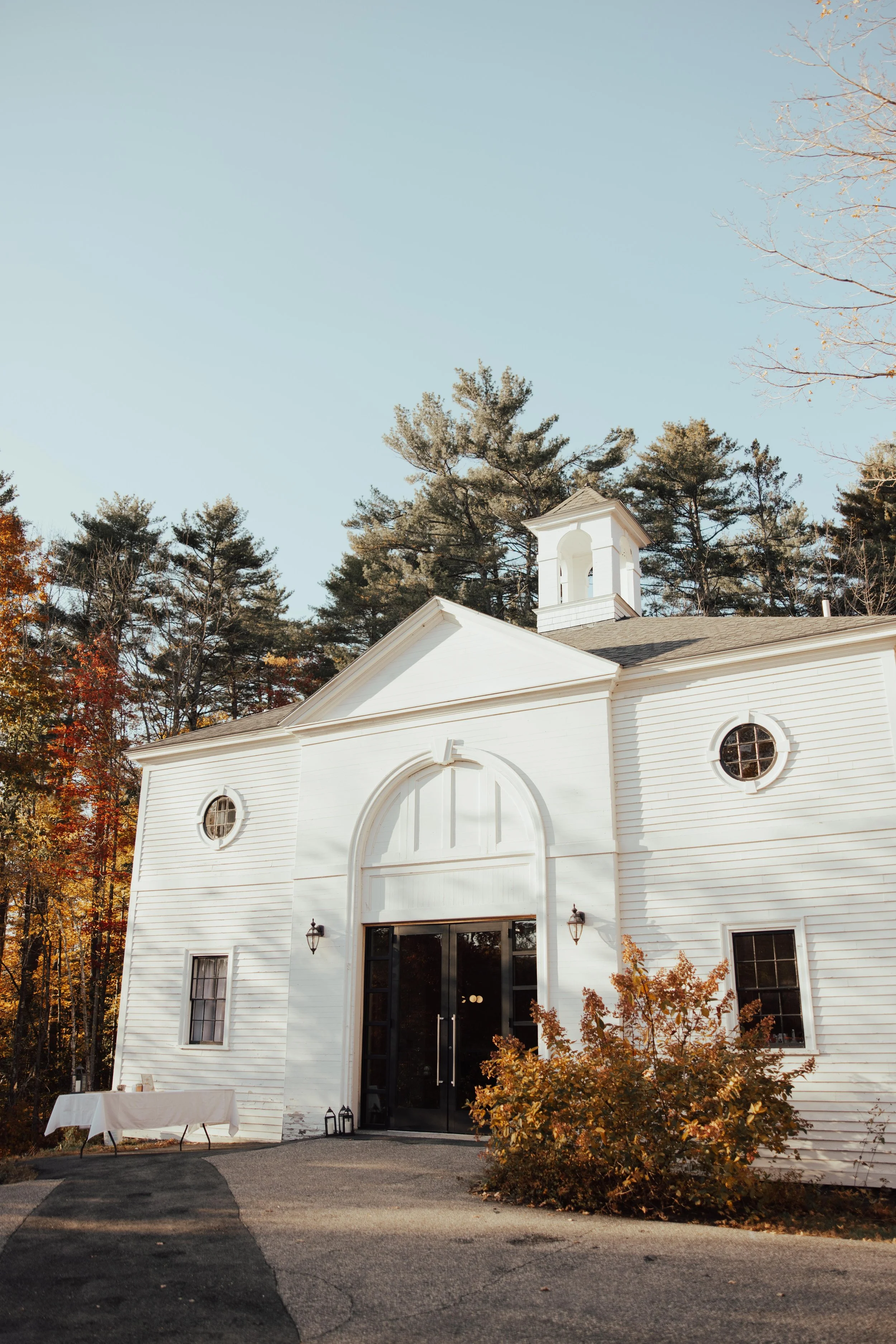 White church with black doors, surrounded by trees with autumn leaves, and a table with a white cloth outside.
