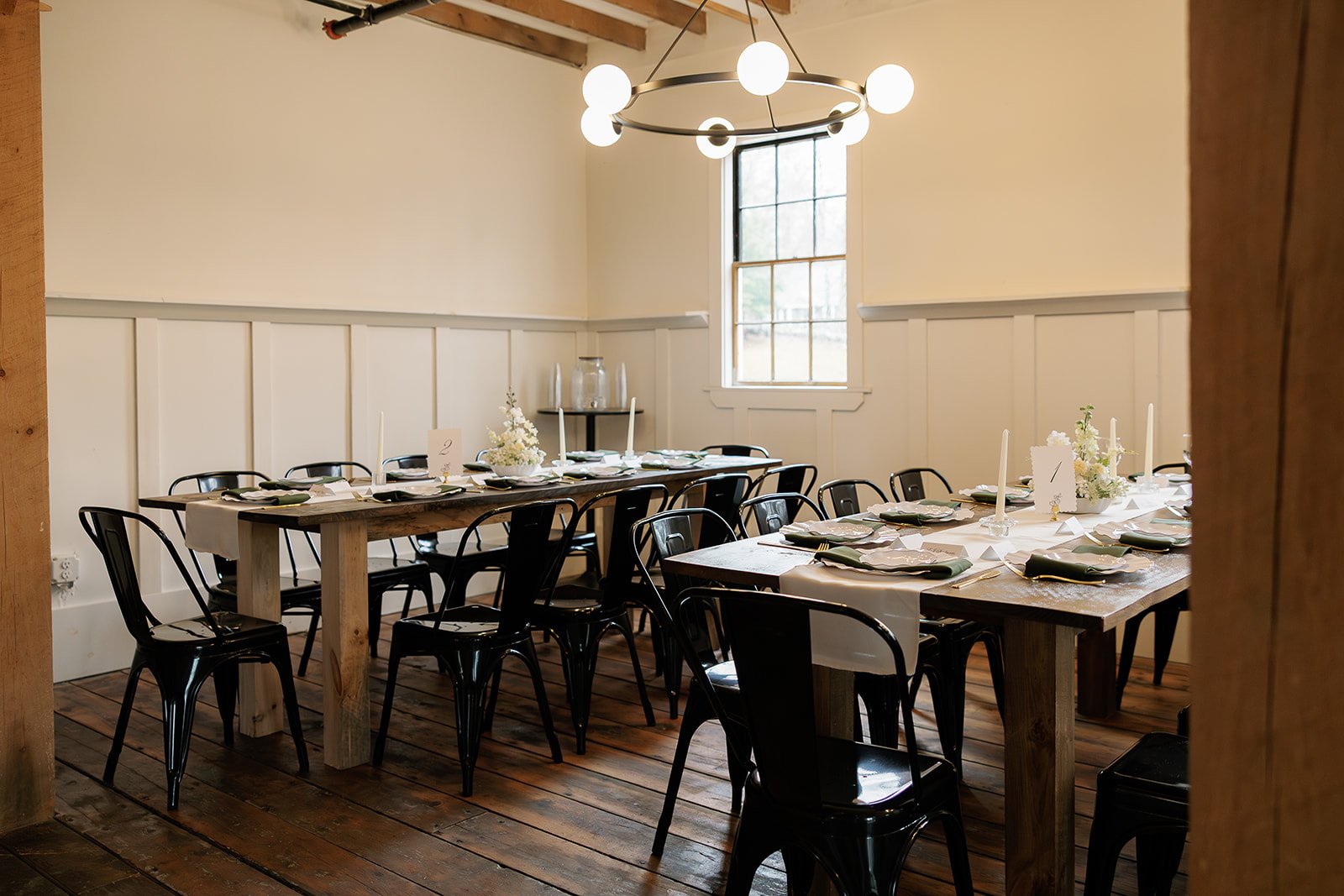 Empty dining room with two wooden tables set for a meal, black chairs, white napkins, candles, and floral centerpieces, with a window and modern chandelier