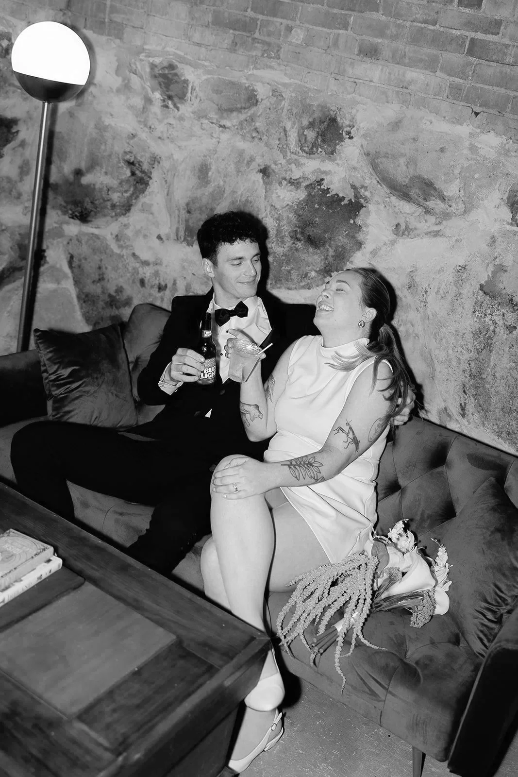 A man in formal attire and a woman in a sleeveless dress are sitting on a sofa, smiling and holding drinks, in the cozy speakeasy setting of our Norway Maine wedding estate.