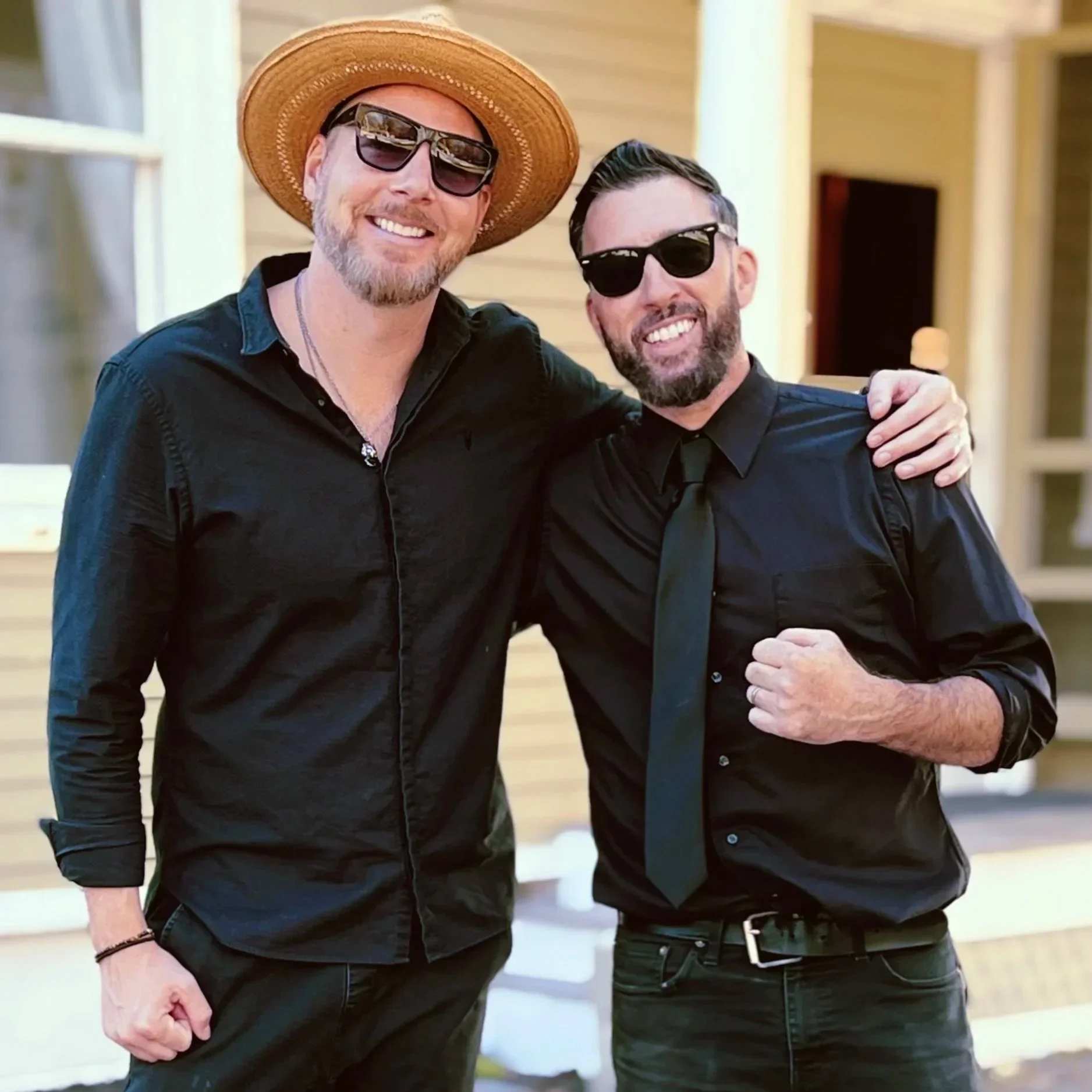 Two men wearing black shirts and sunglasses, smiling and posing with arms around each other outdoors on a sunny wedding in Norway Maine