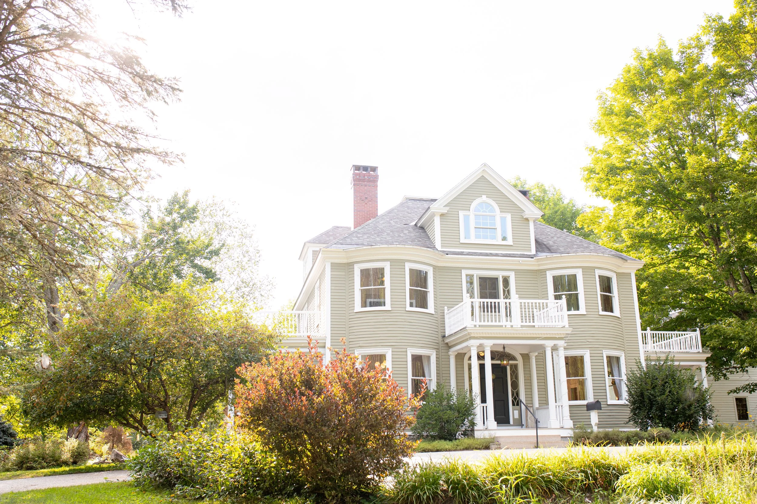 Front view of a large, light green Victorian house with white trim, multiple windows, a small balcony, and a chimney, surrounded by lush green trees and bushes under bright sunlight.