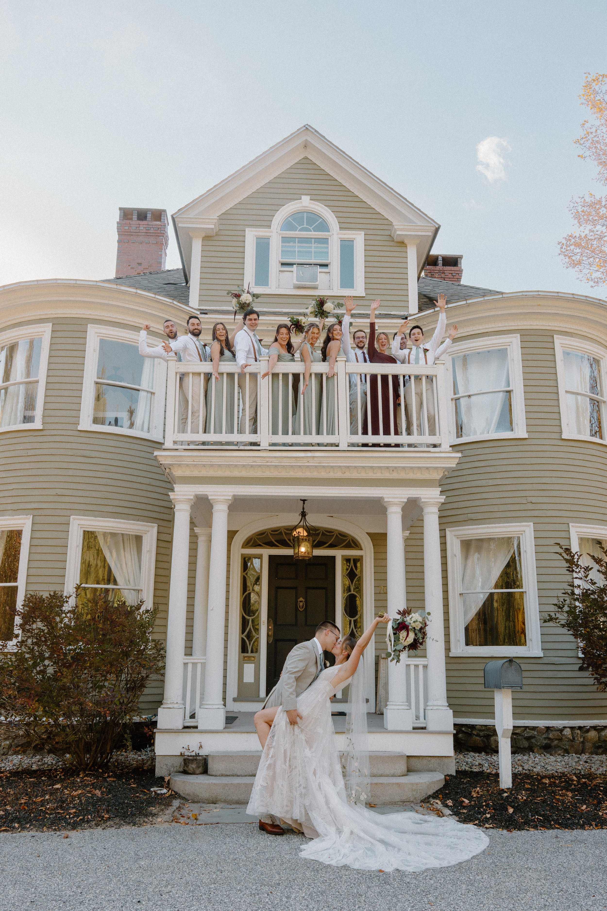 Wedding celebration at a large house with bride and groom kissing in front, wedding party on the balcony above celebrating, bride holding bouquet, groom lifting bride, autumn trees in the background.