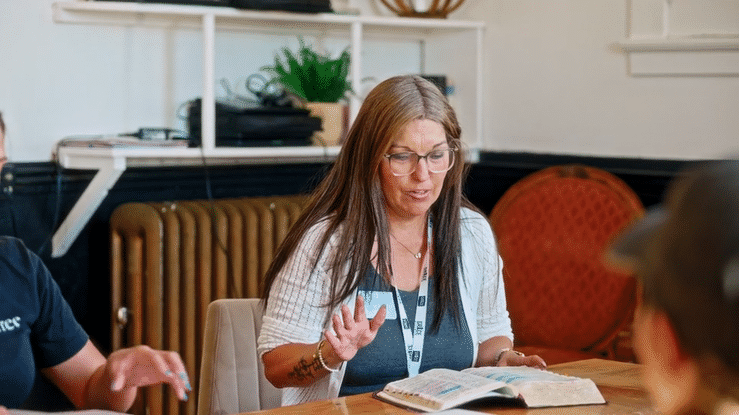 A woman with long brown hair, glasses, and a white cardigan gestures while speaking at a table during a meeting or discussion, with books and other people visible around her in a room with bookshelves and a radiator.