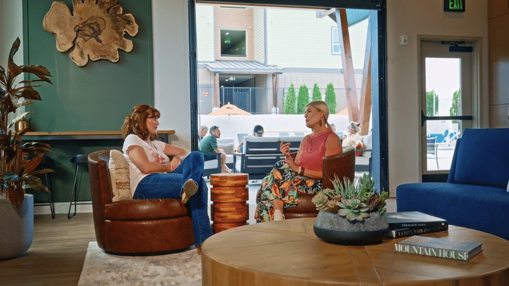 Two women sitting and talking inside a cozy café or lounge area with large windows, tabletop plants, and modern decor.