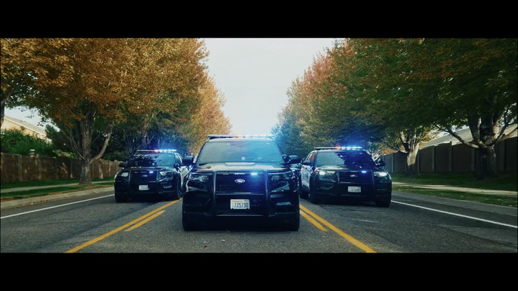 Kennewick Police Department's latest recruiting video. Three police cars with flashing lights blocking a residential street lined with fall-colored trees. Police officers marching.