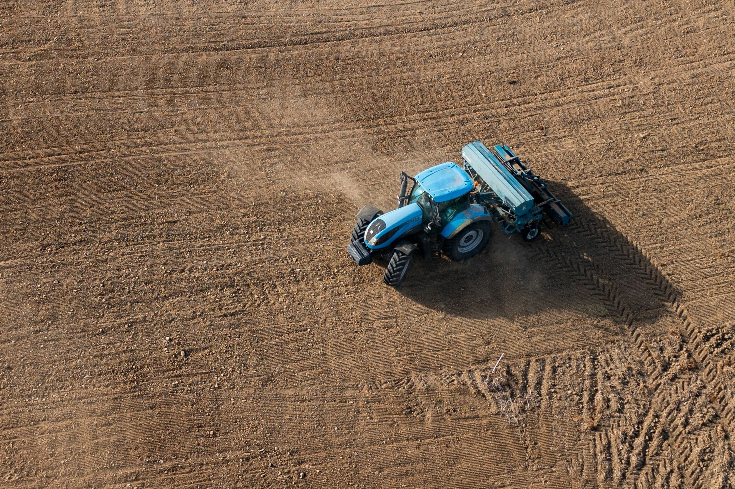 Aerial view of a blue tractor working on a large brown field, creating tire tracks in the soil.