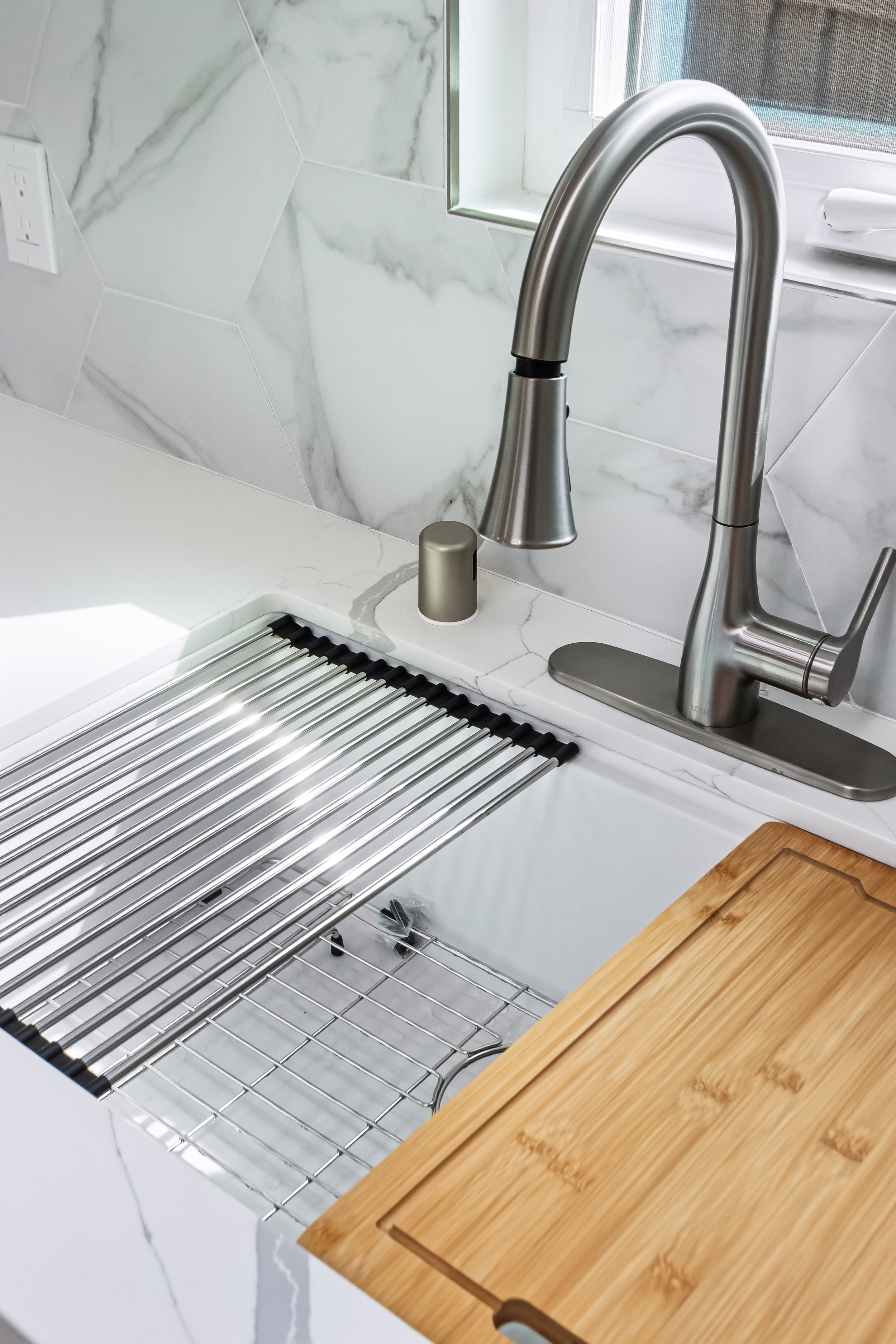 A modern kitchen sink with a stainless steel faucet, a wooden cutting board, a dish drying rack, and marble backsplash tiles.