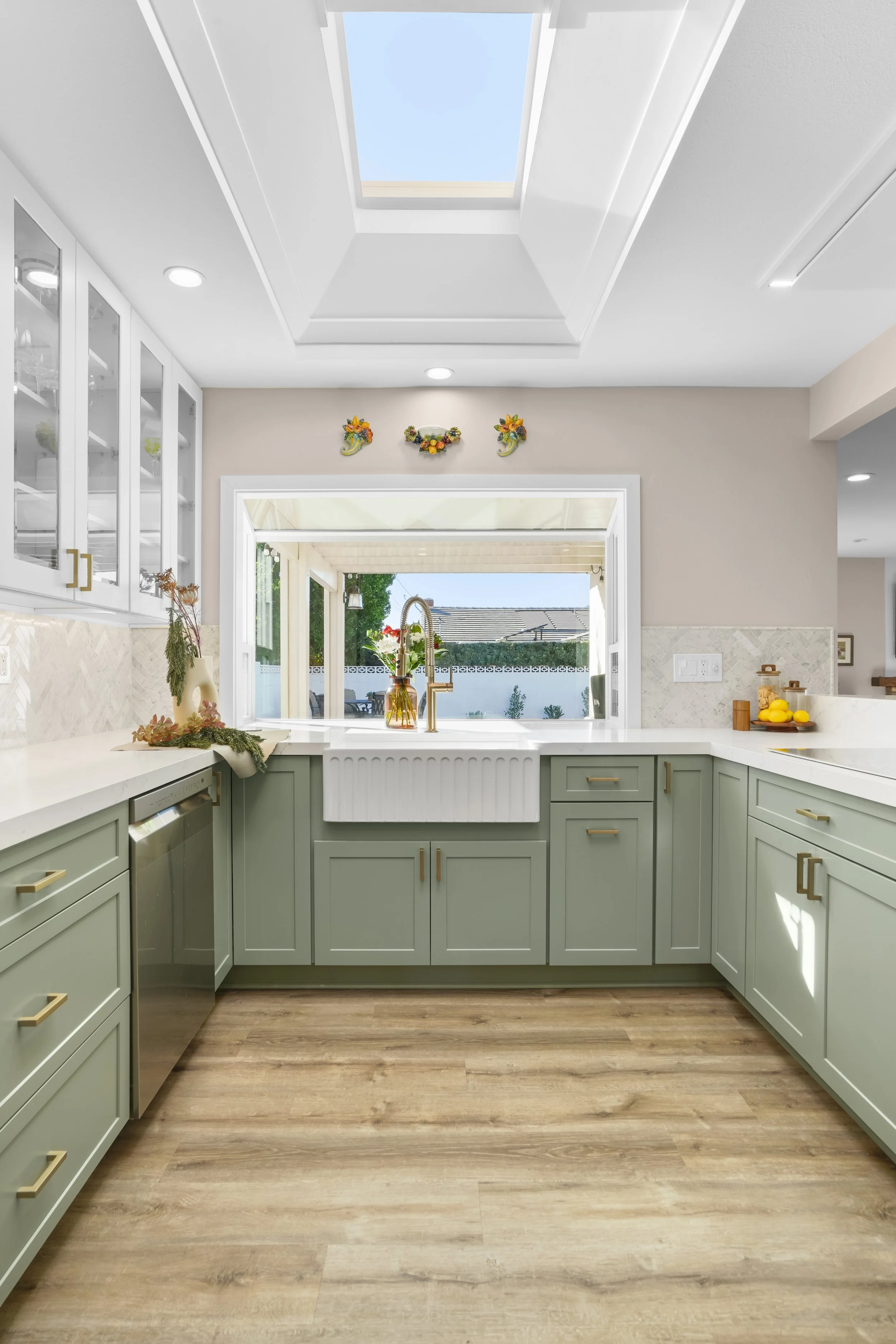 A bright kitchen in Orange County with a skylight, light green cabinets, a white farmhouse sink, and a window overlooking a backyard. Decor includes a vase with flowers and a bowl of lemons.