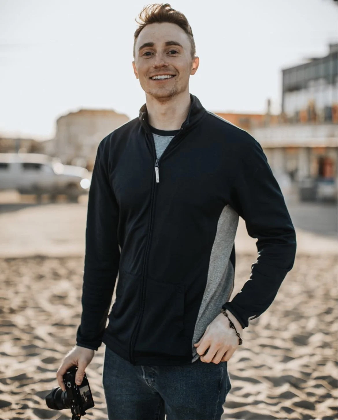 Matt Collamer smiling outdoors on a sunny day, holding a camera, standing on the beach.