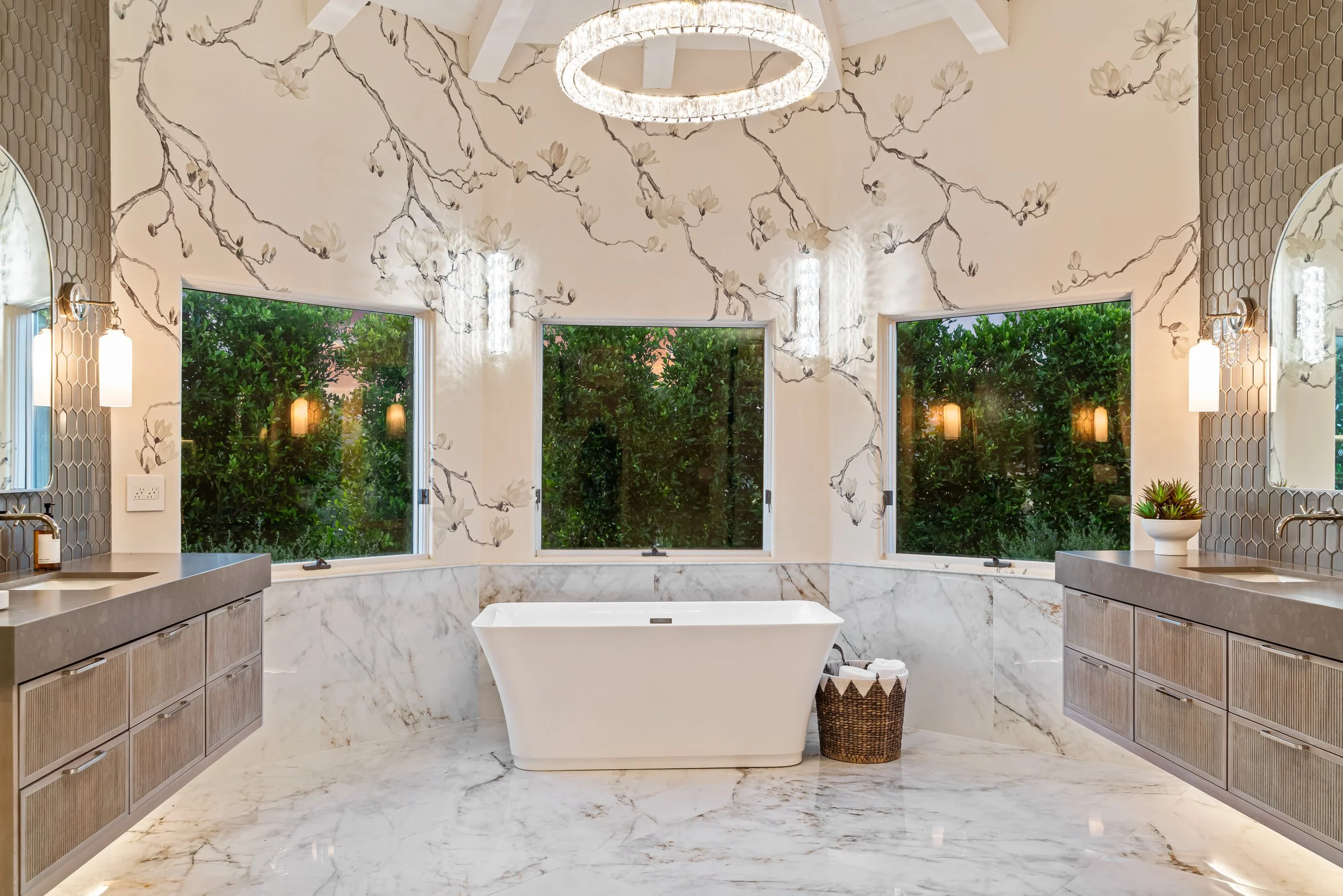Luxurious Rancho Santa Fe bathroom with a freestanding bathtub in front of three large windows, marble flooring, and wall with floral mural. There are two sinks with gray countertops, mirrored cabinets, and decorative lighting.