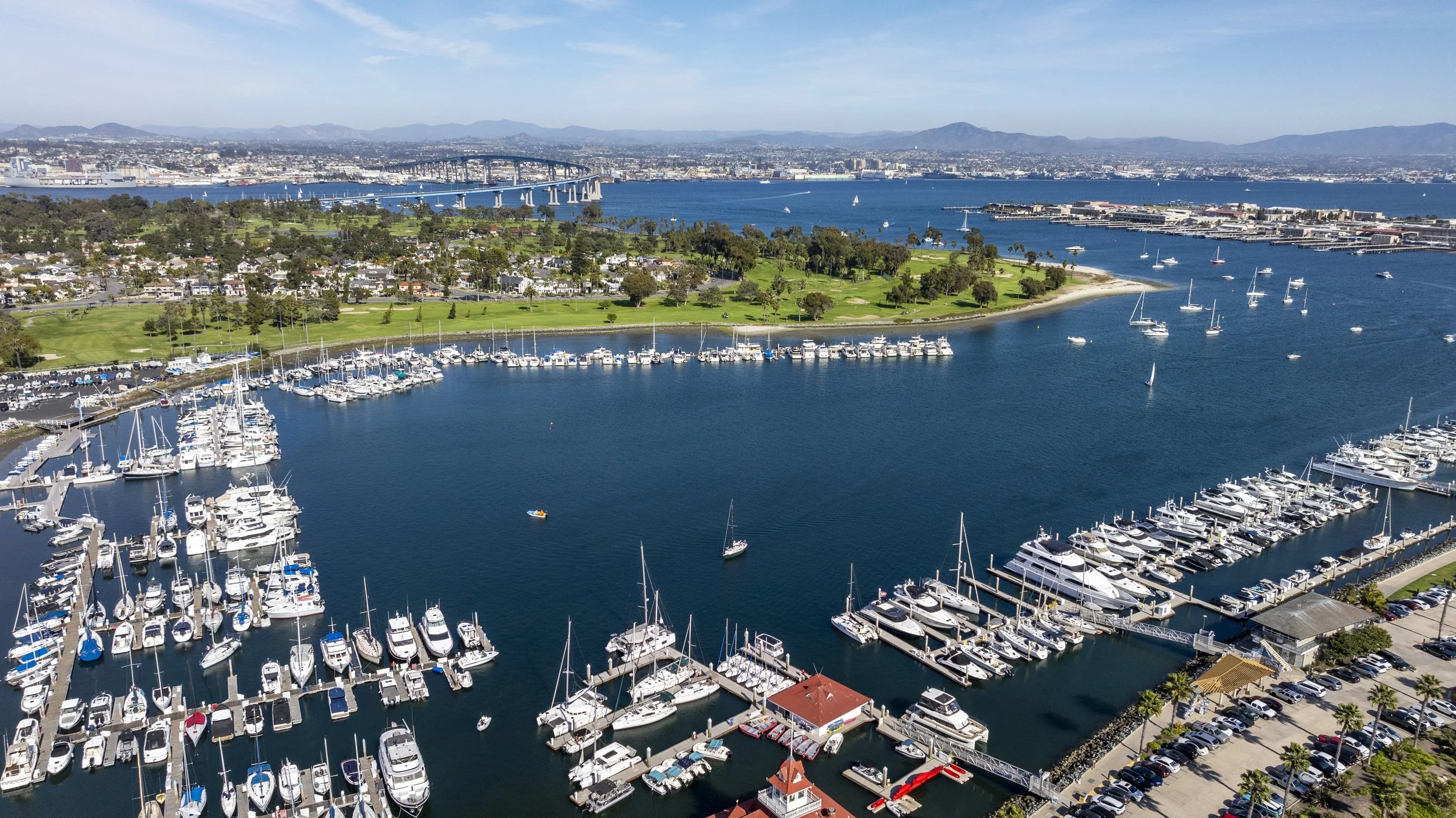 Drone photo of San Diego marina with numerous sailboats and yachts docked and a cityscape in the background, including the Coronado bridge crossing a large body of water under a clear blue sky.