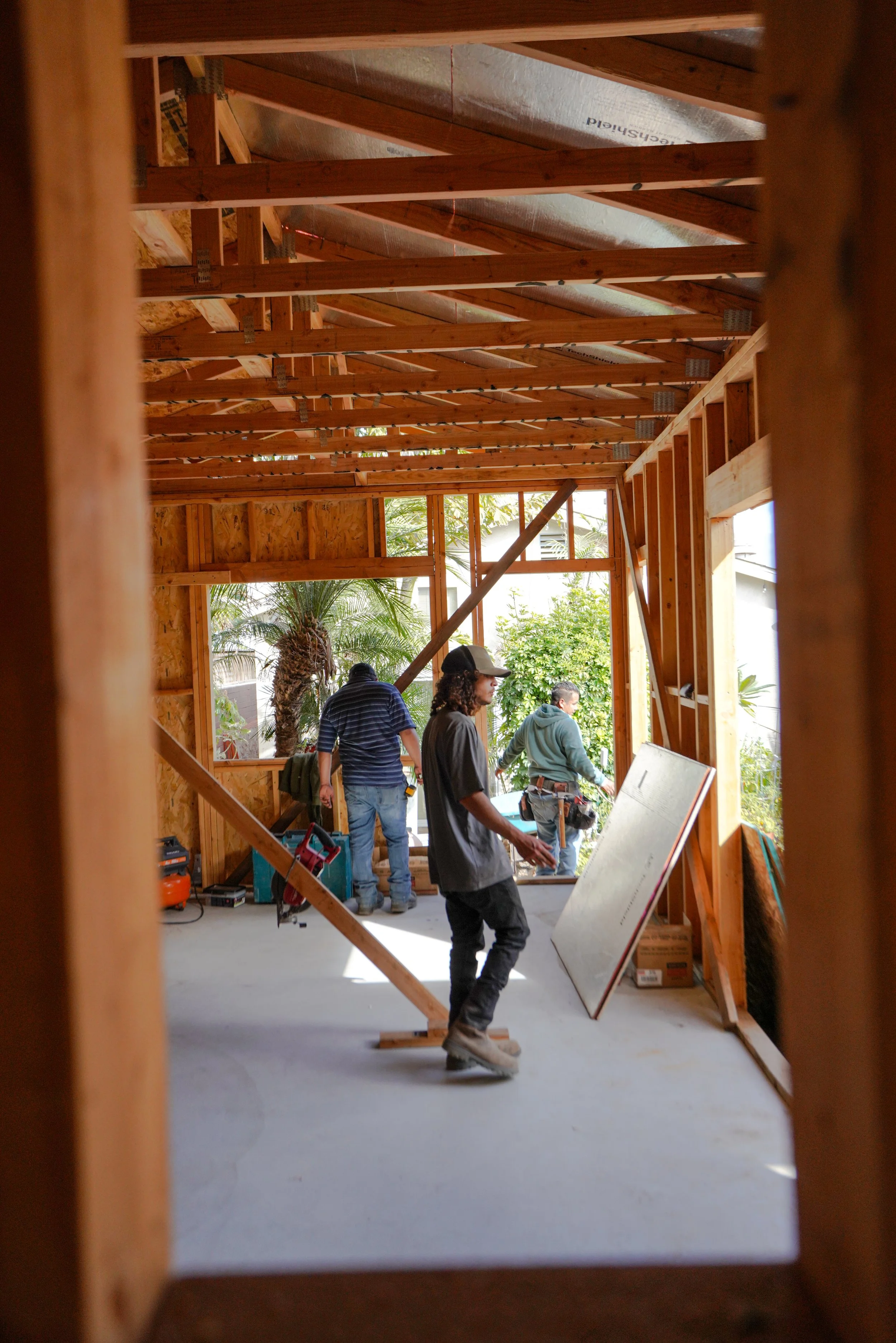 San Diego construction workers building a house frame inside a partially completed wooden structure with visible roof trusses and large window openings, with outdoor greenery in the background.