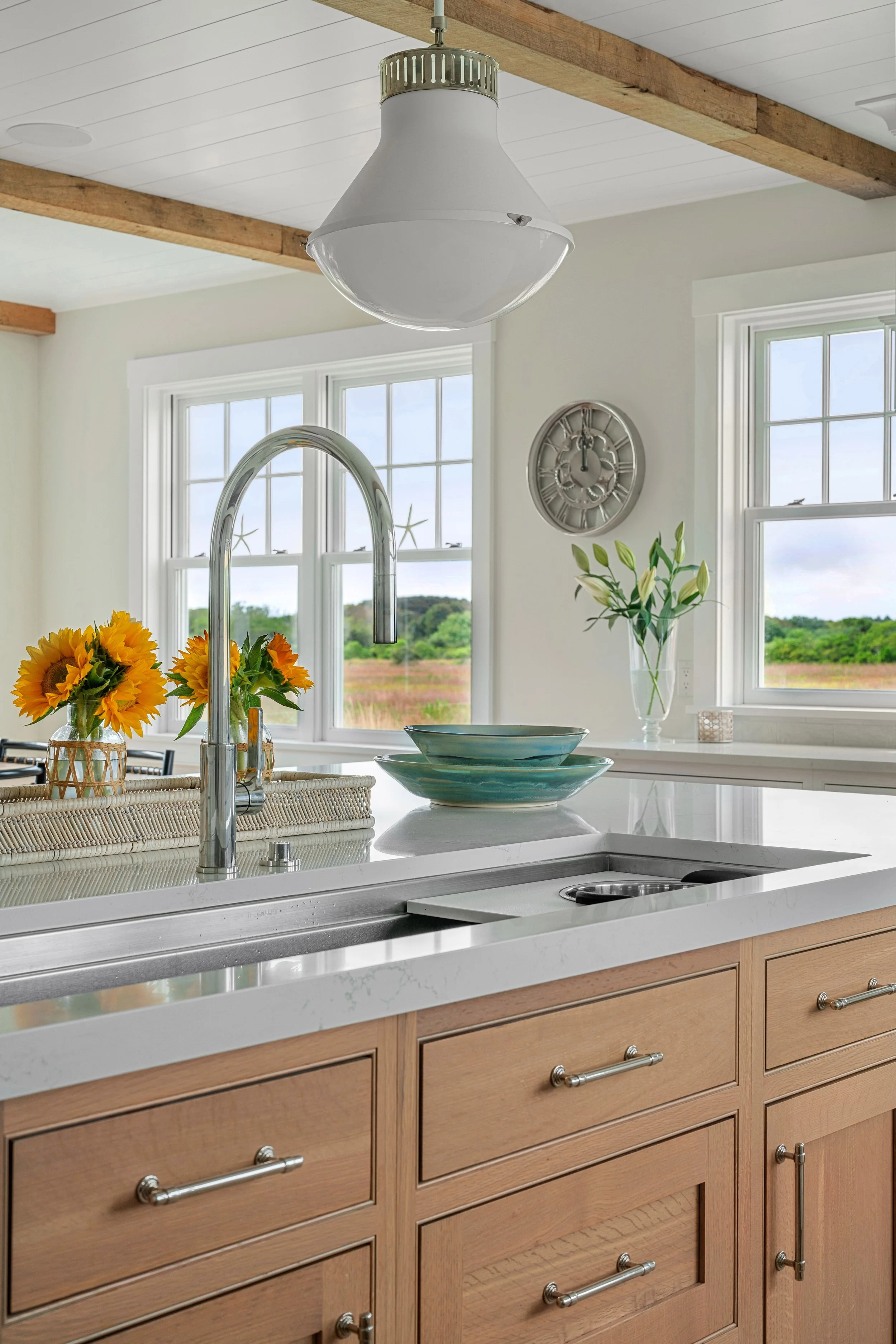 Kitchen with wooden cabinets, marble countertop, a sink with a high-arc chrome faucet, two sunflower vases, and a green decorative bowl, with view of open field through windows in the background.