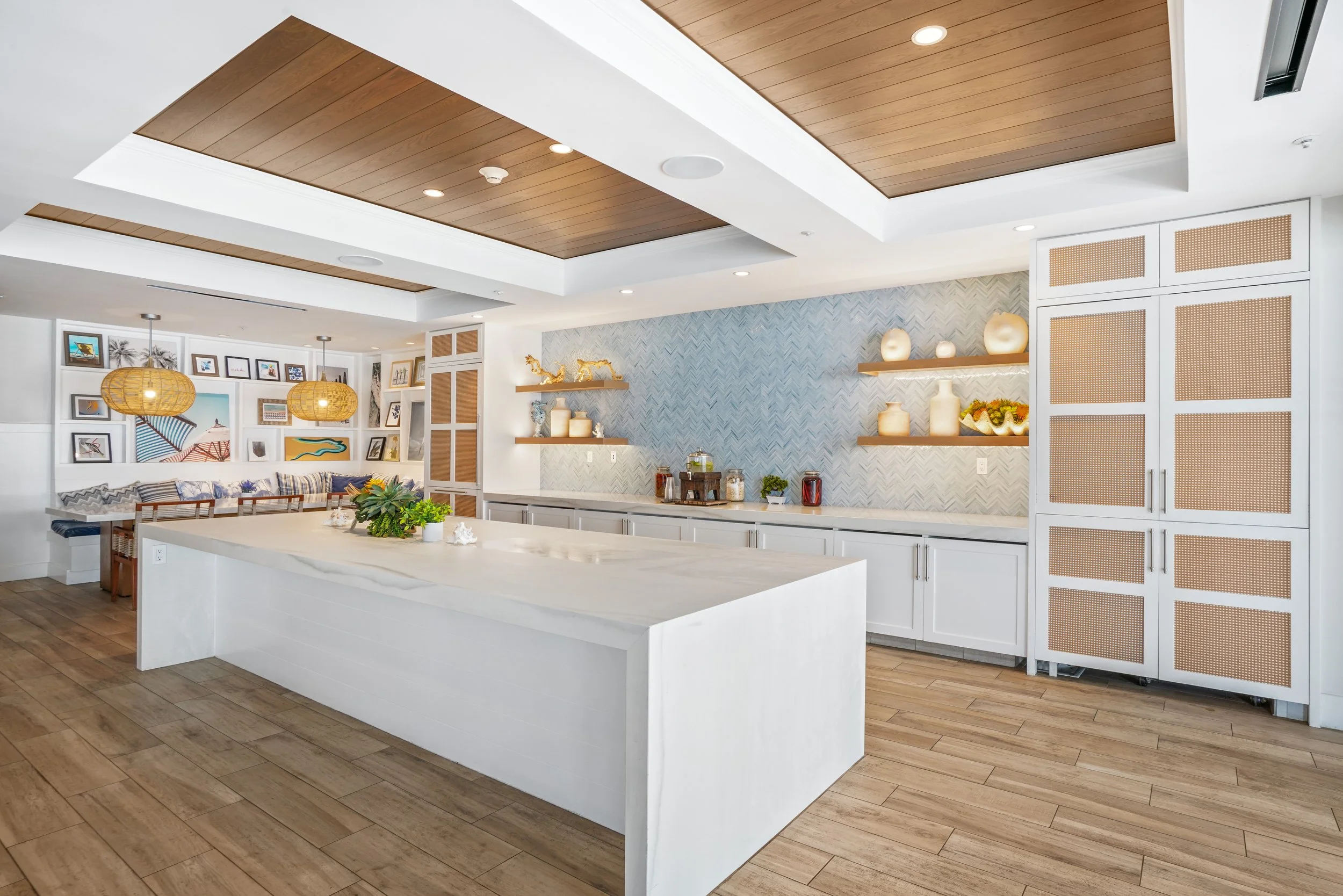 Modern kitchen and dining area at Hotel Del Coronado with white cabinetry, a large island, wooden shelves, and a cozy seating nook with framed artwork and hanging rattan lights.