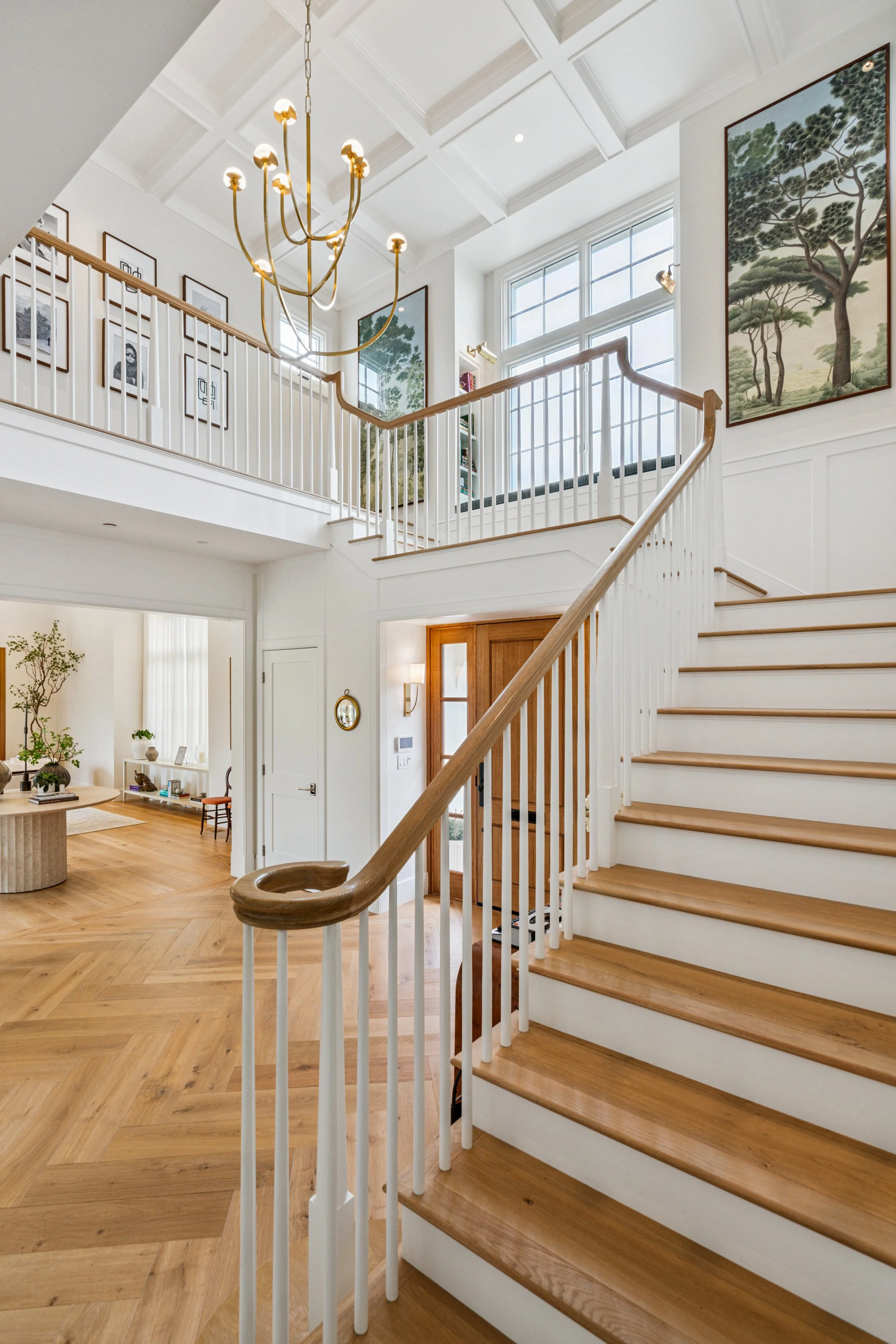 Bright, airy interior of a two-story home with a wooden staircase, large windows, and art on the walls. a chandelier hangs from the ceiling, and the space features white walls and wooden flooring.