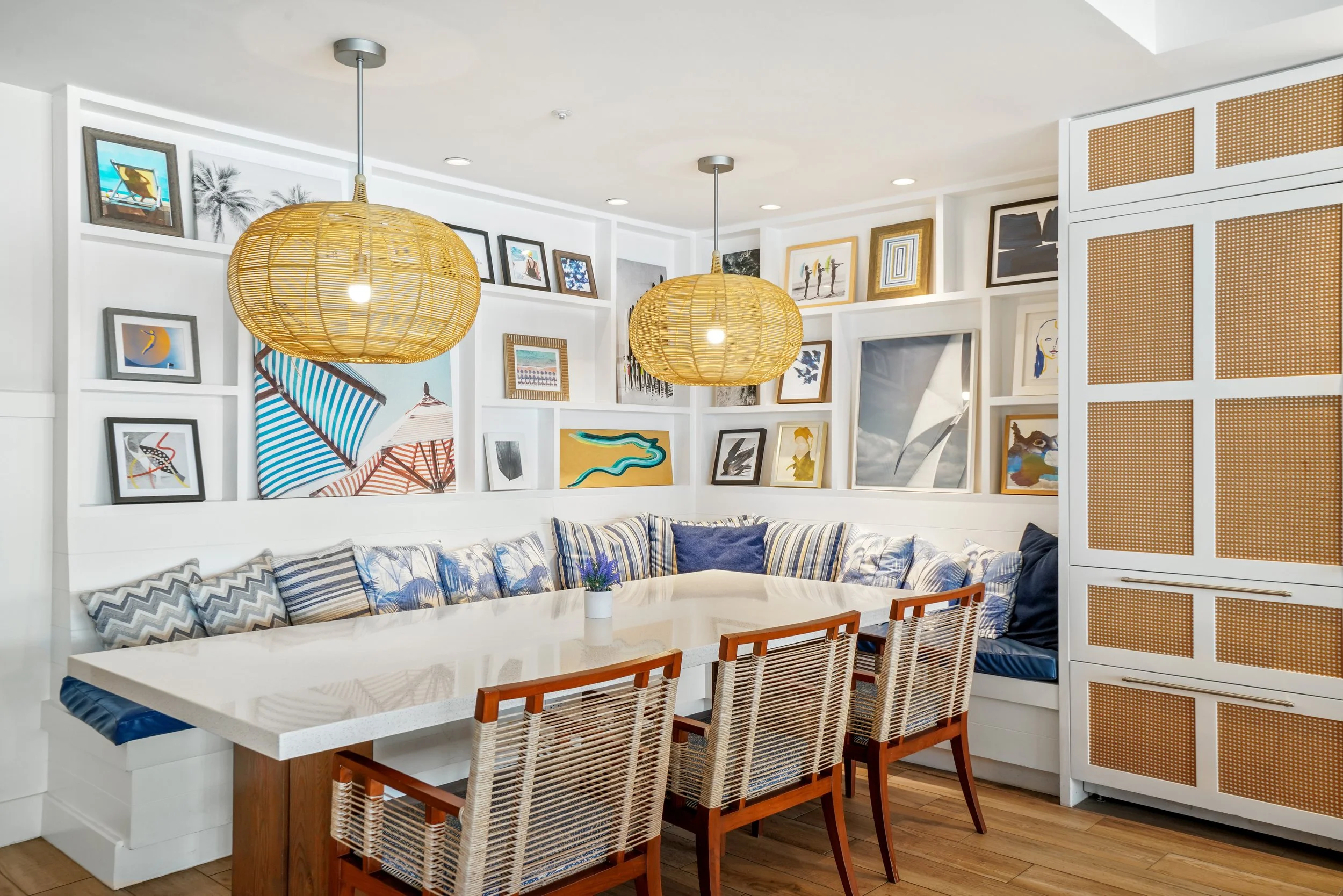 A luxury dining area with a white table, rattan chairs, blue and white cushions, a built-in corner bench, hanging woven pendant lights, and a wall decorated with an eclectic collection of framed artwork at Hotel Del Coronado.