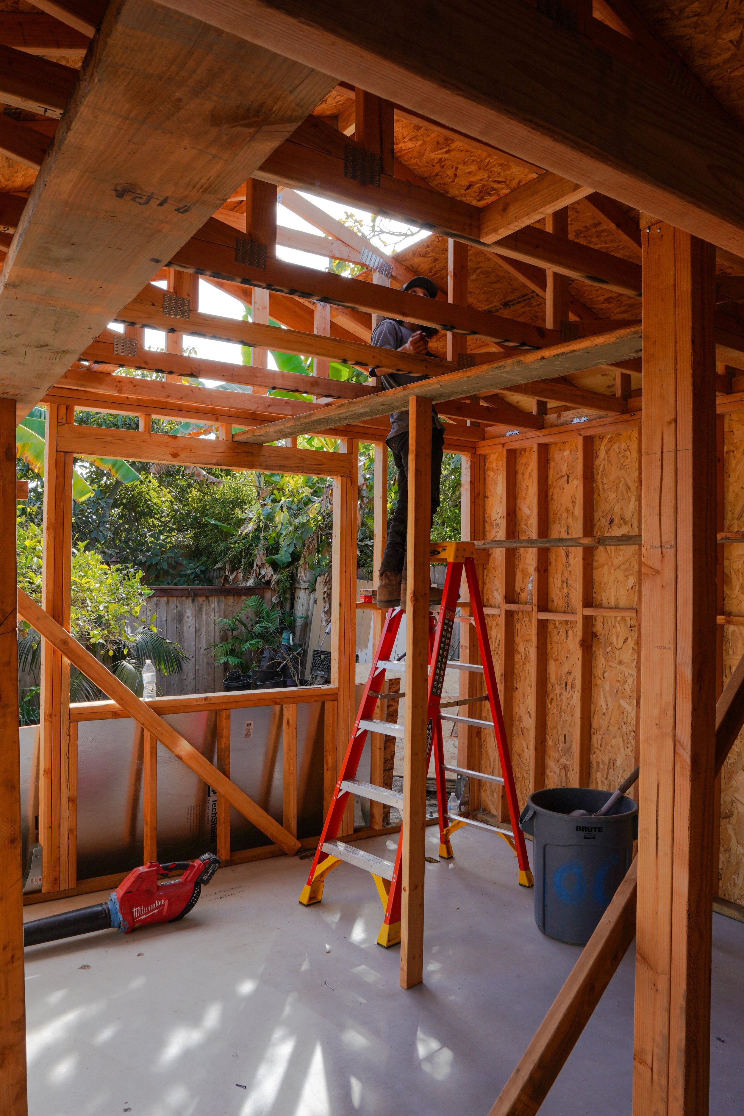 Behind the scenes photo of the interior of a house under construction in San Diego with wooden framing, a worker on a stepladder, and construction tools.